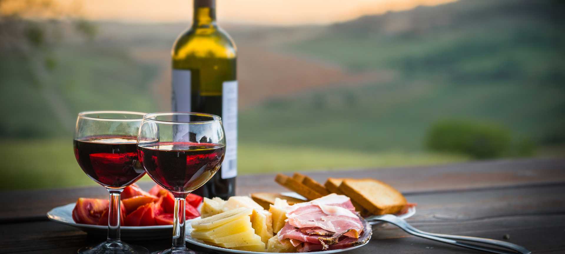 Two glasses of red wine are served alongside plates of bread, cheese, cold cuts and tomatoes, with a countryside view behind