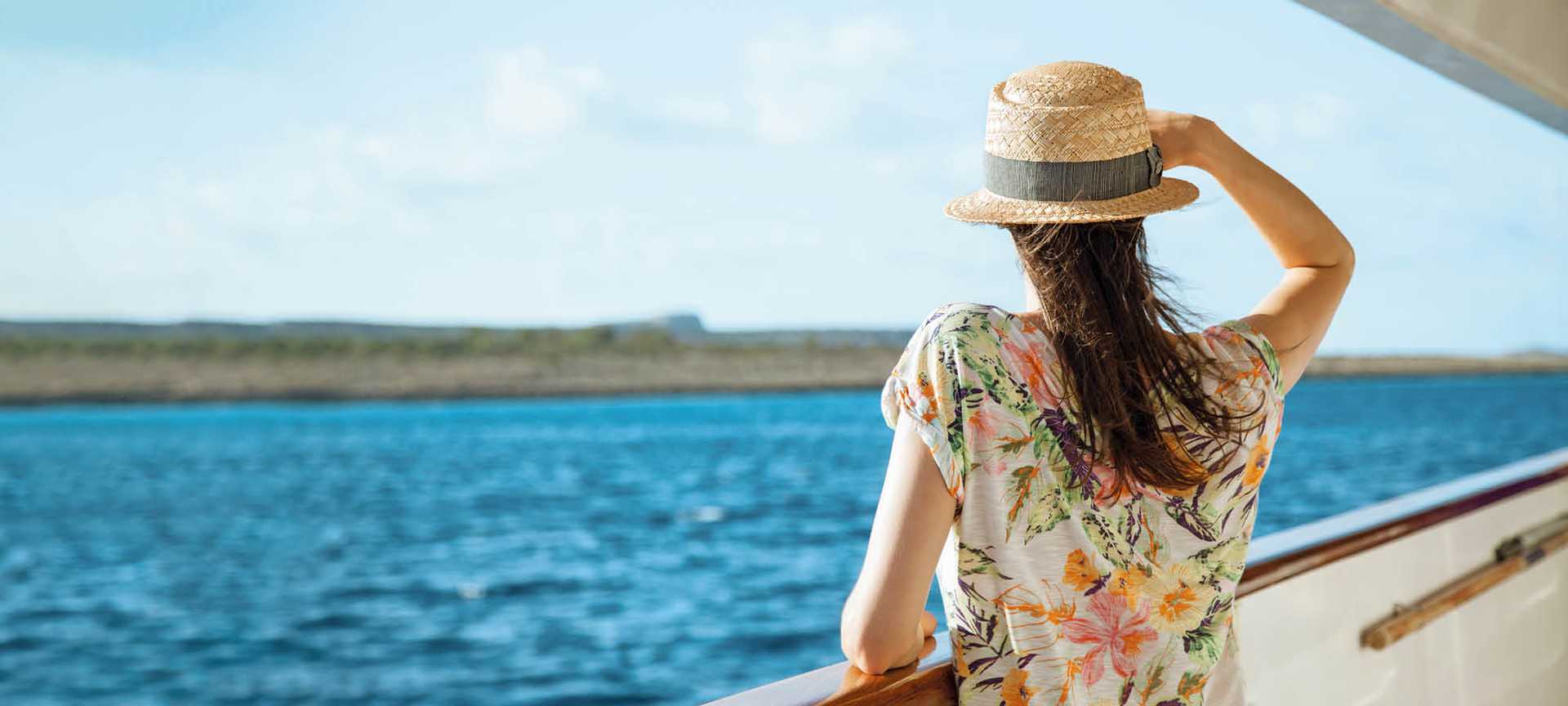 A woman in a sun hat stands on a cruise ship looking out to sea