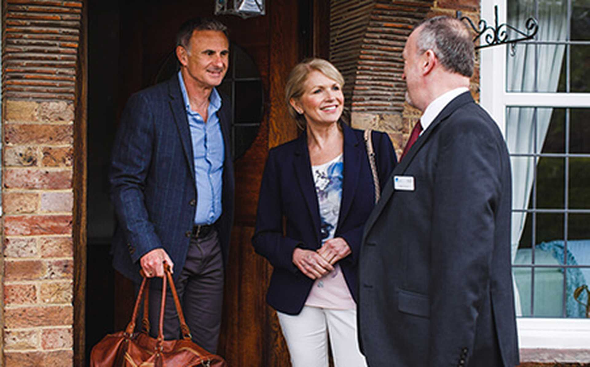 Titan VIP door-to-door travel service chauffeur greeting a man and woman at their front door. They are smiling and the man has a brown leather holdall.