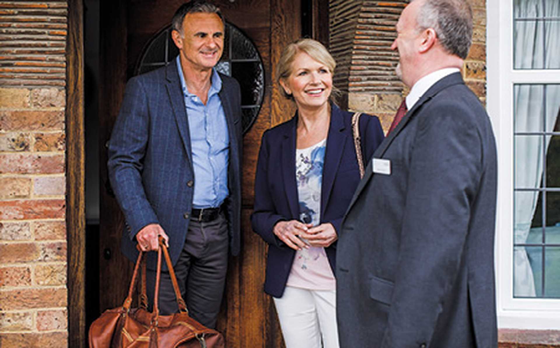 Titan VIP door-to-door travel service chauffeur greeting a man and woman at their front door. They are smiling and the man has a brown leather holdall.