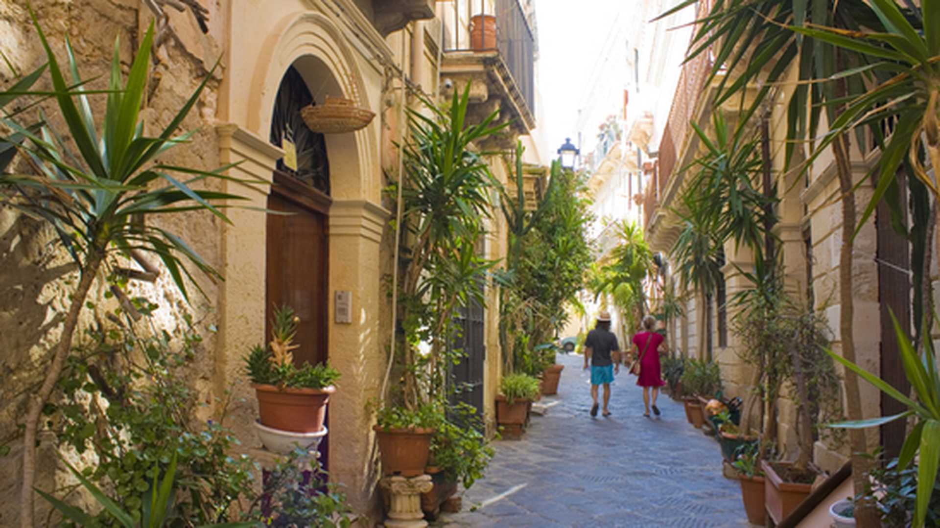 Two people walk down a narrow lane lined with plants and sandstone townhouses in Ortigia, the old town of Syracuse, Sicily