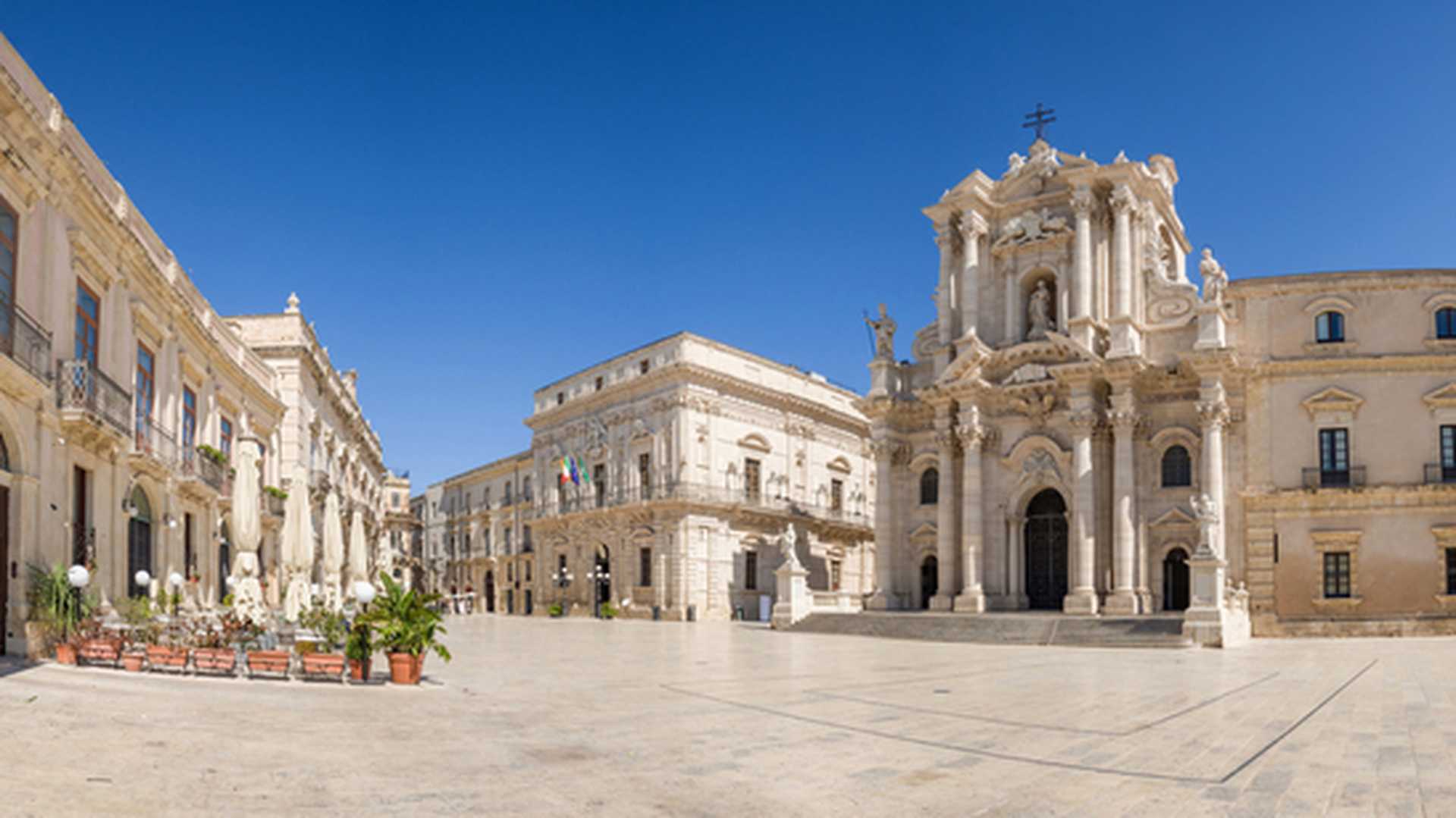 Restaurant tables set out beneath white parasols in a sunny piazza, with Ortigia's baroque cathedral behind