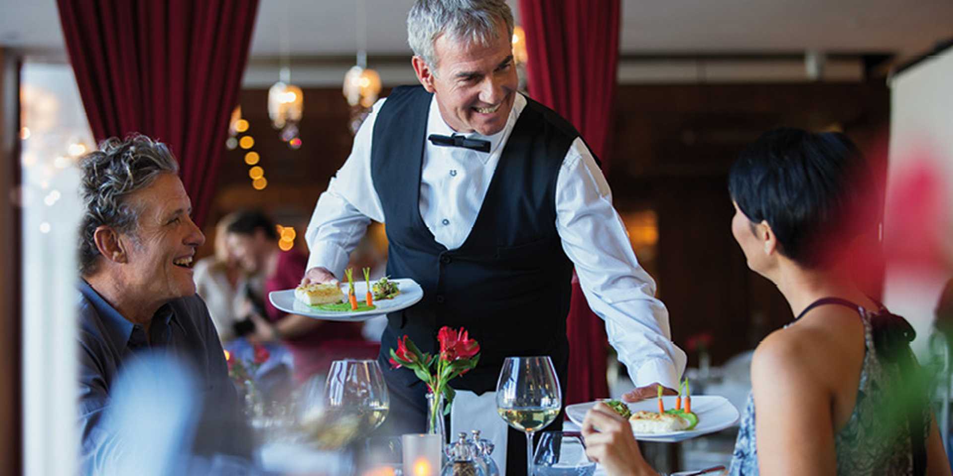 Smiling waiter serving fancy dishes to mature couple sitting at table in restaurant