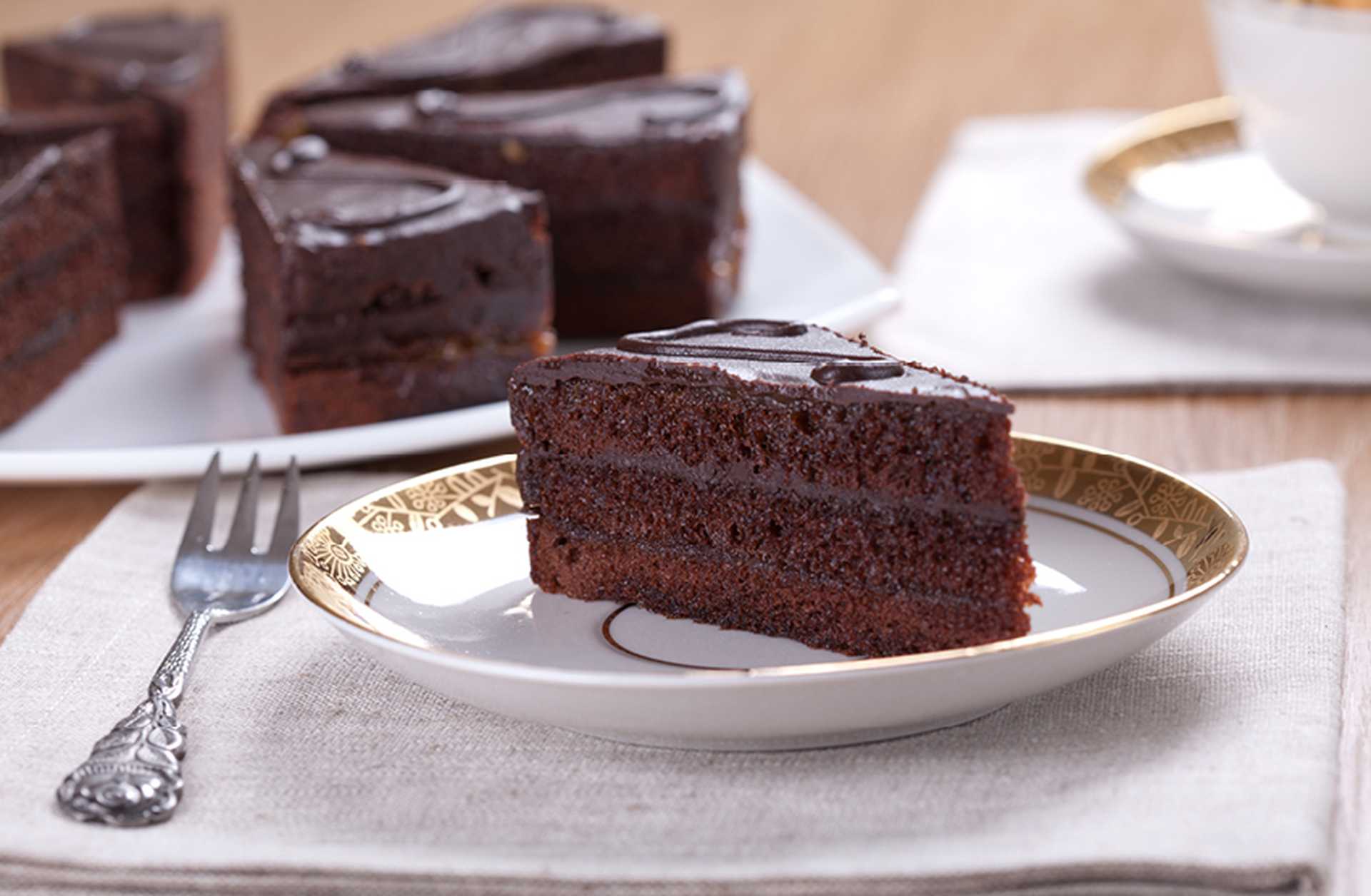 A slice of sachertorte is served on a white plate with a small fork, with more slices visible in the background