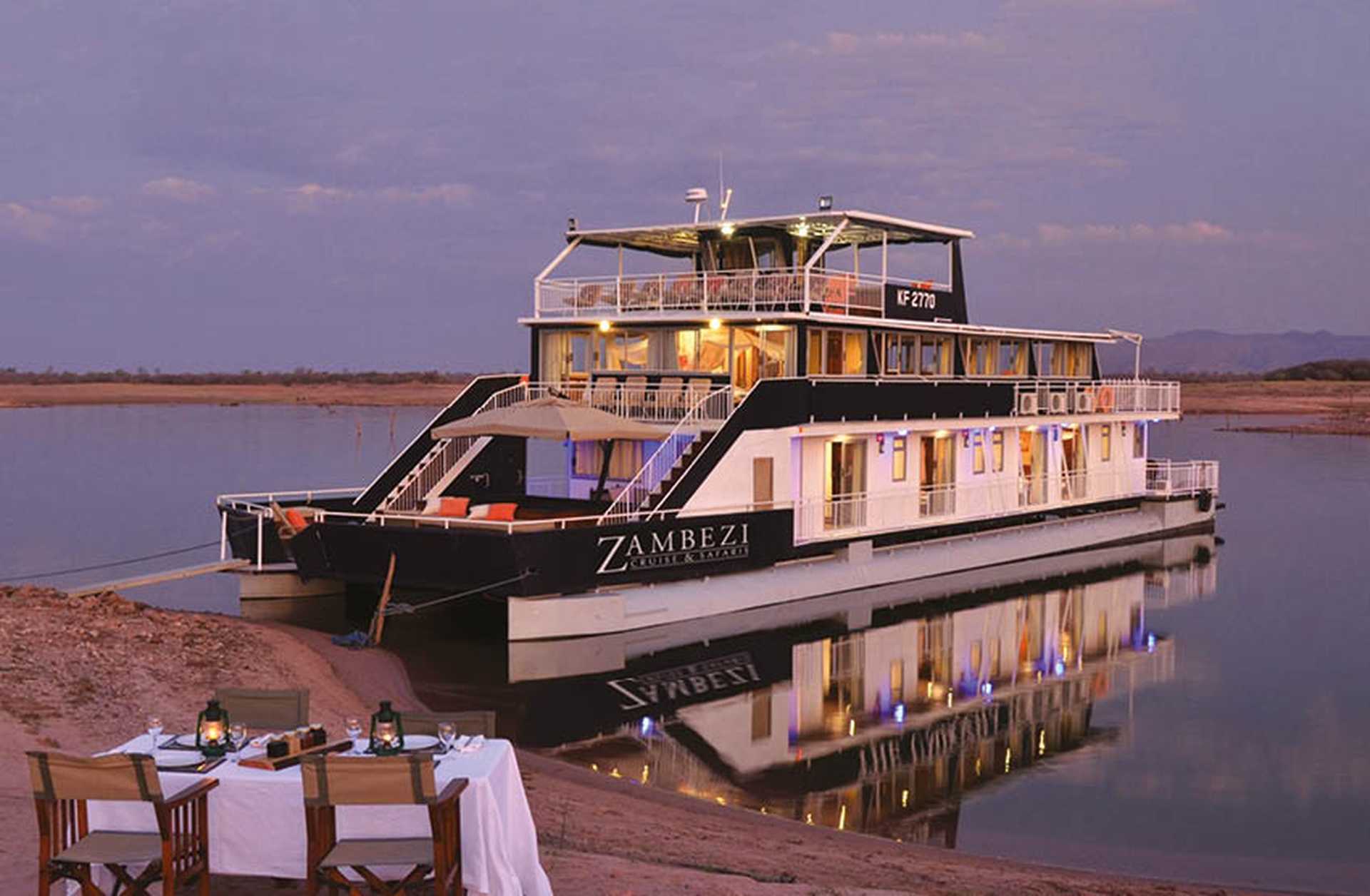A dining table and chairs are set up beside the water next to a Zambezi Cruise Safari boat 