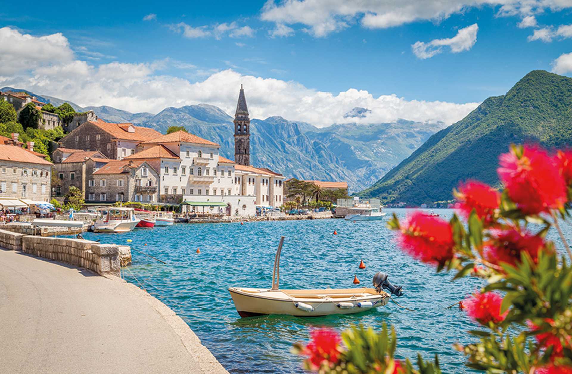 The historic town of Perast on the Bay of Kotor, with red flowers in the foreground and mountains in the background