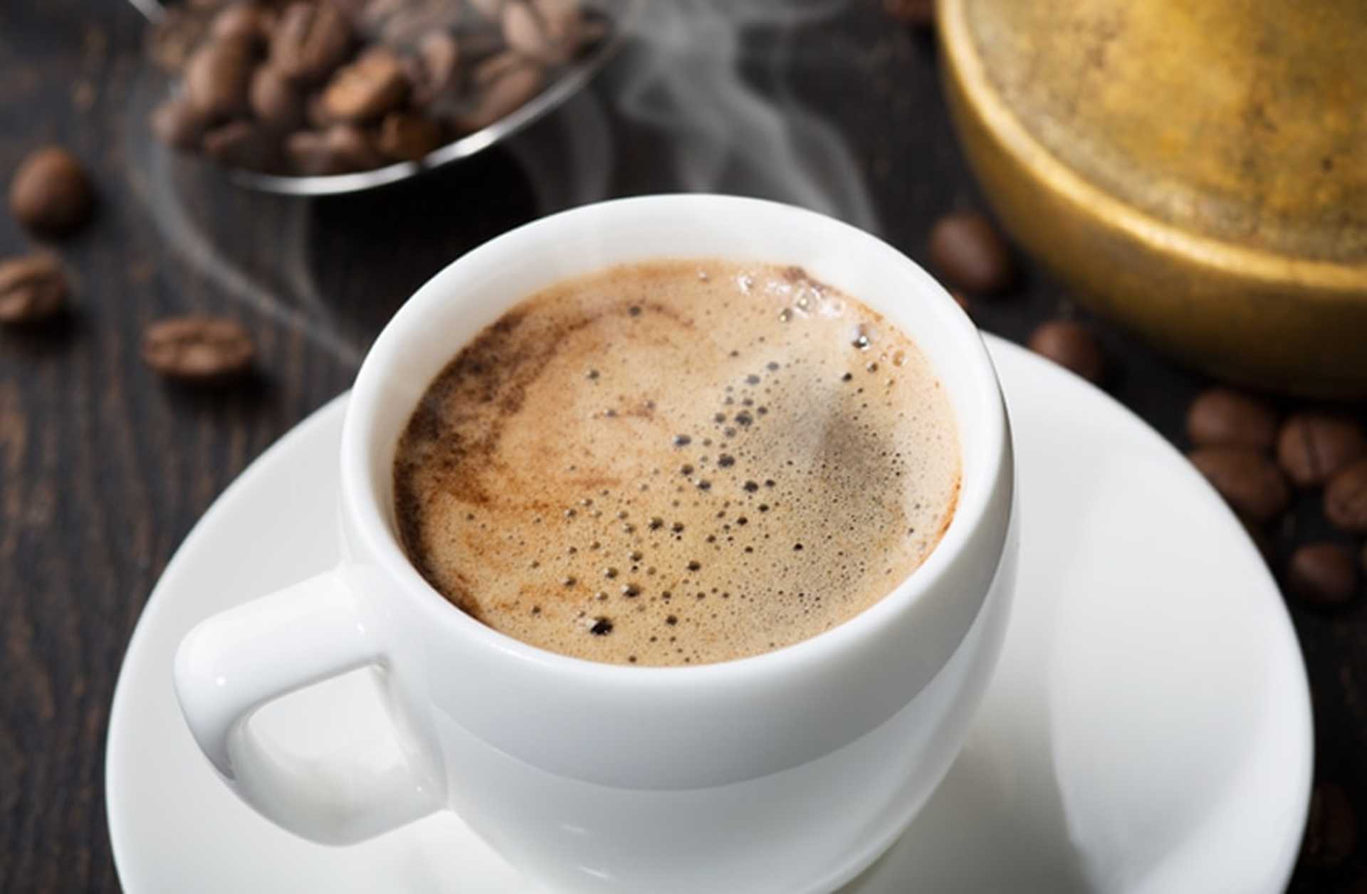 Steam rises from a cup of coffee, with a silver dish of coffee beans behind