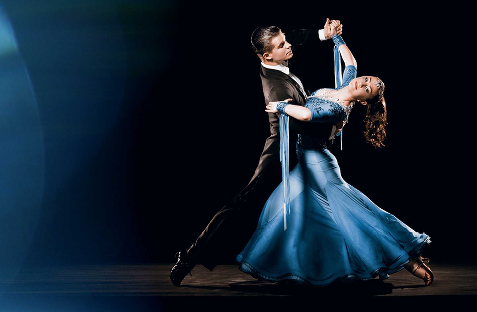 A man in a black suit and a woman in a blue ballgown dance the waltz