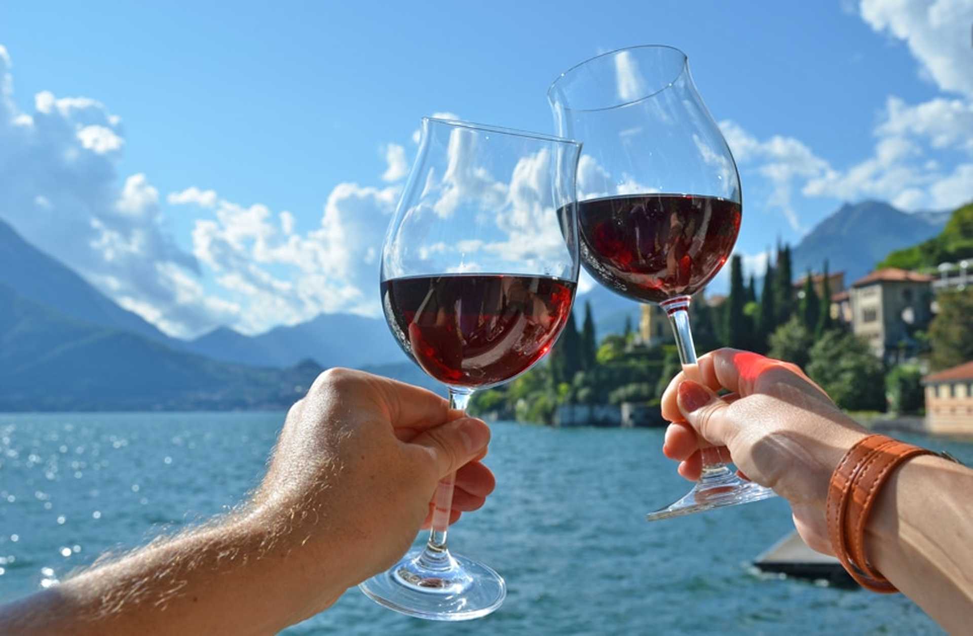Two people clink glasses of red wine together with Lake Como in the background