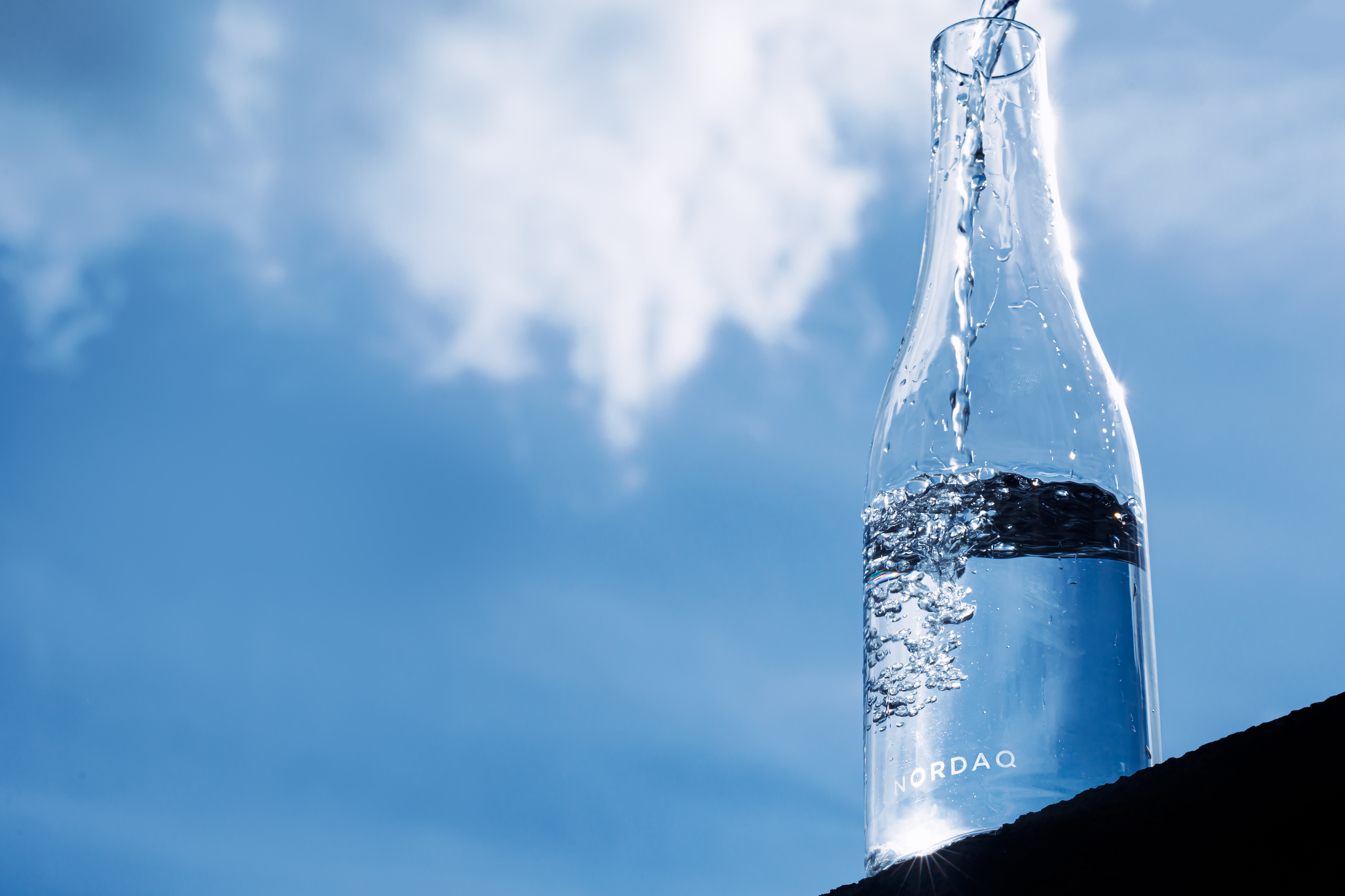 Clear water pours into a glass bottle labeled "Nordaq" against a blue sky with clouds.