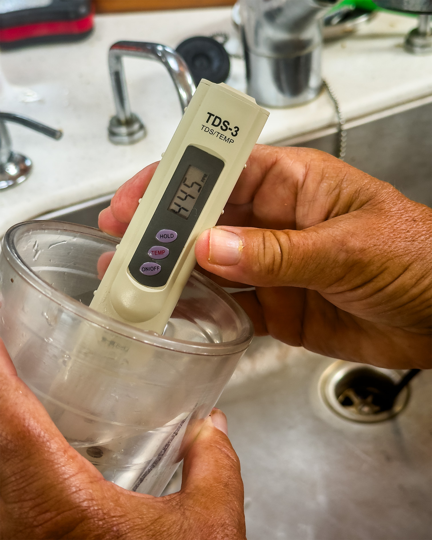 Person measuring water quality with a TDS meter in a glass over a sink, showing a reading of 554 ppm on the display.