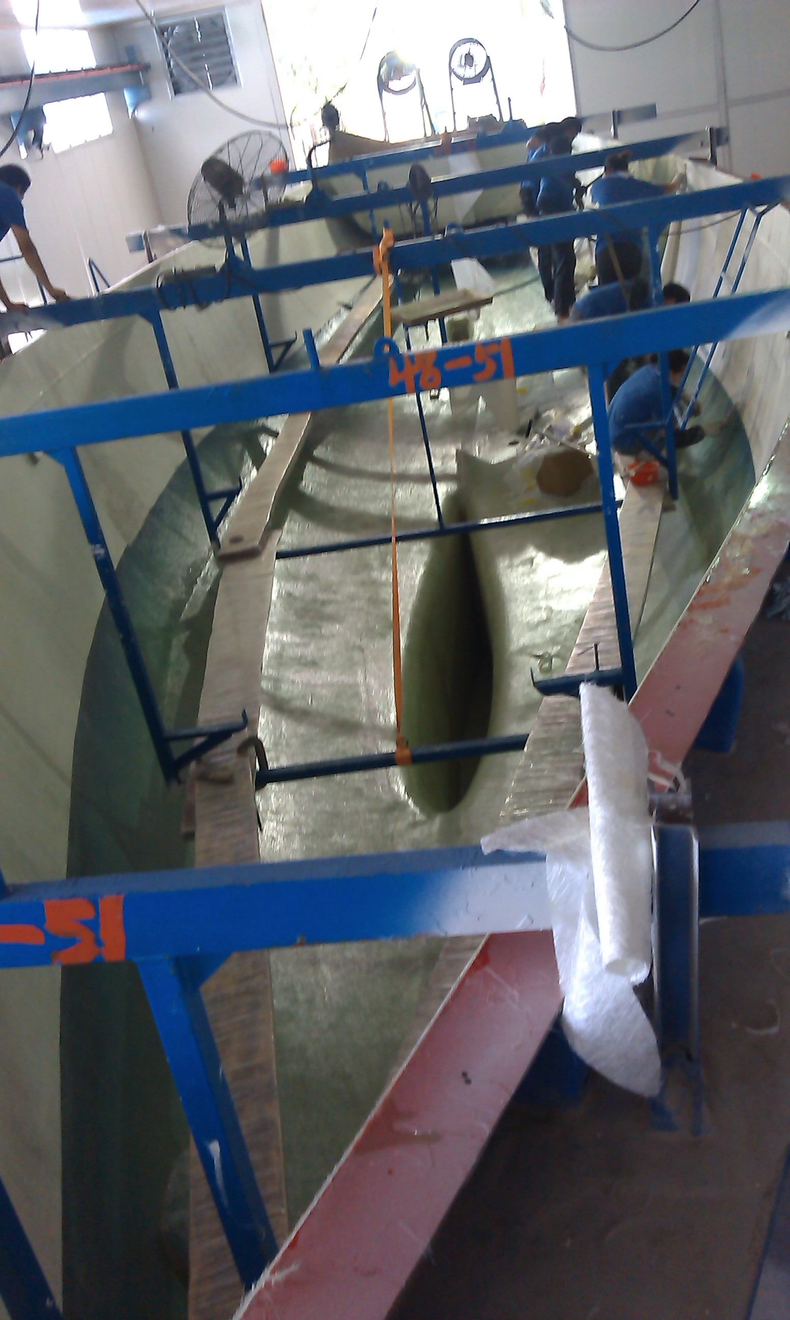 SV Wanderlust's hull under construction in a workshop with blue metal supports, wooden planks, and tools scattered around.