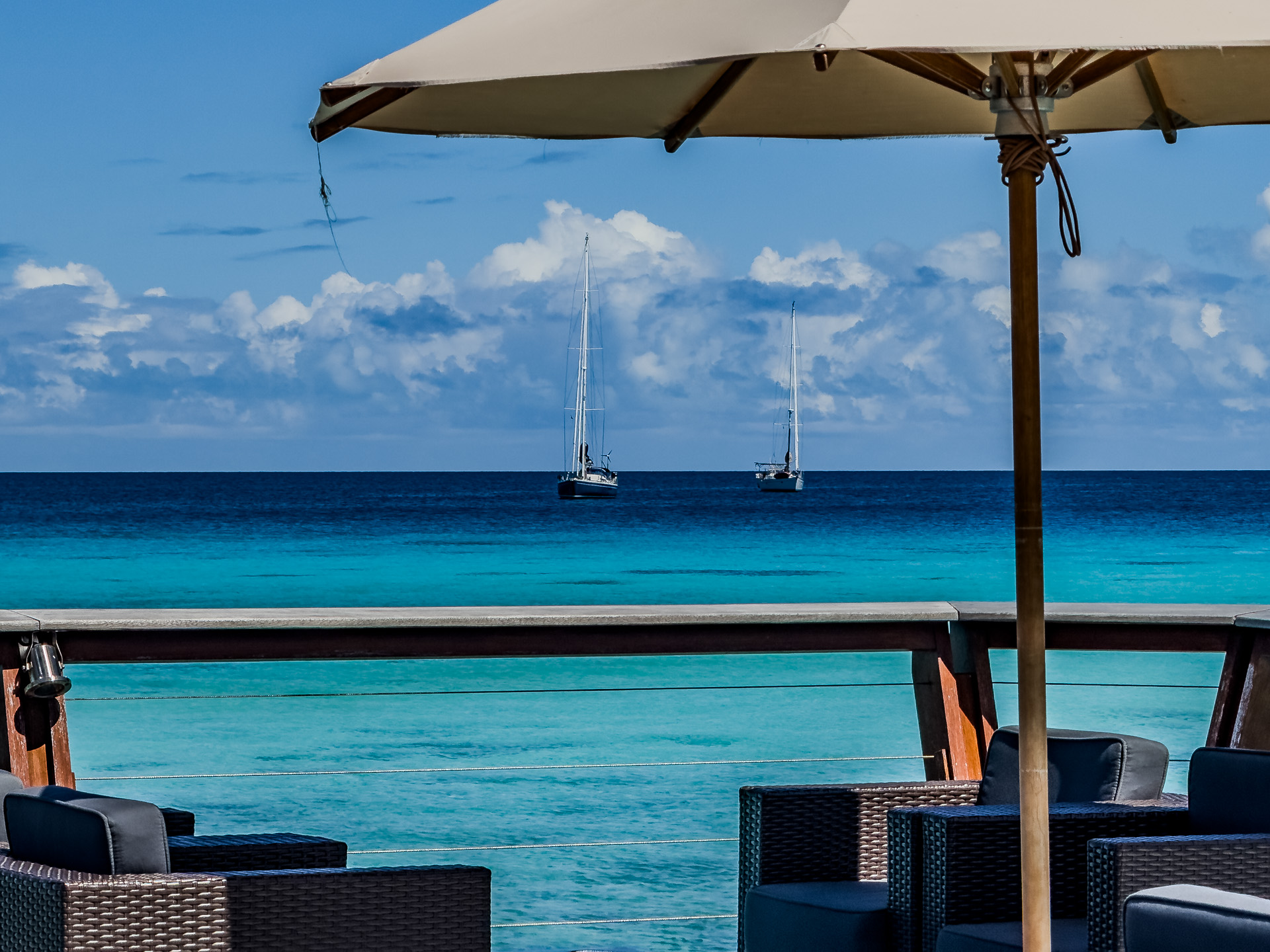 Outdoor seating under an umbrella overlooks a turquoise sea with two sailboats in the distance and a partly cloudy sky.