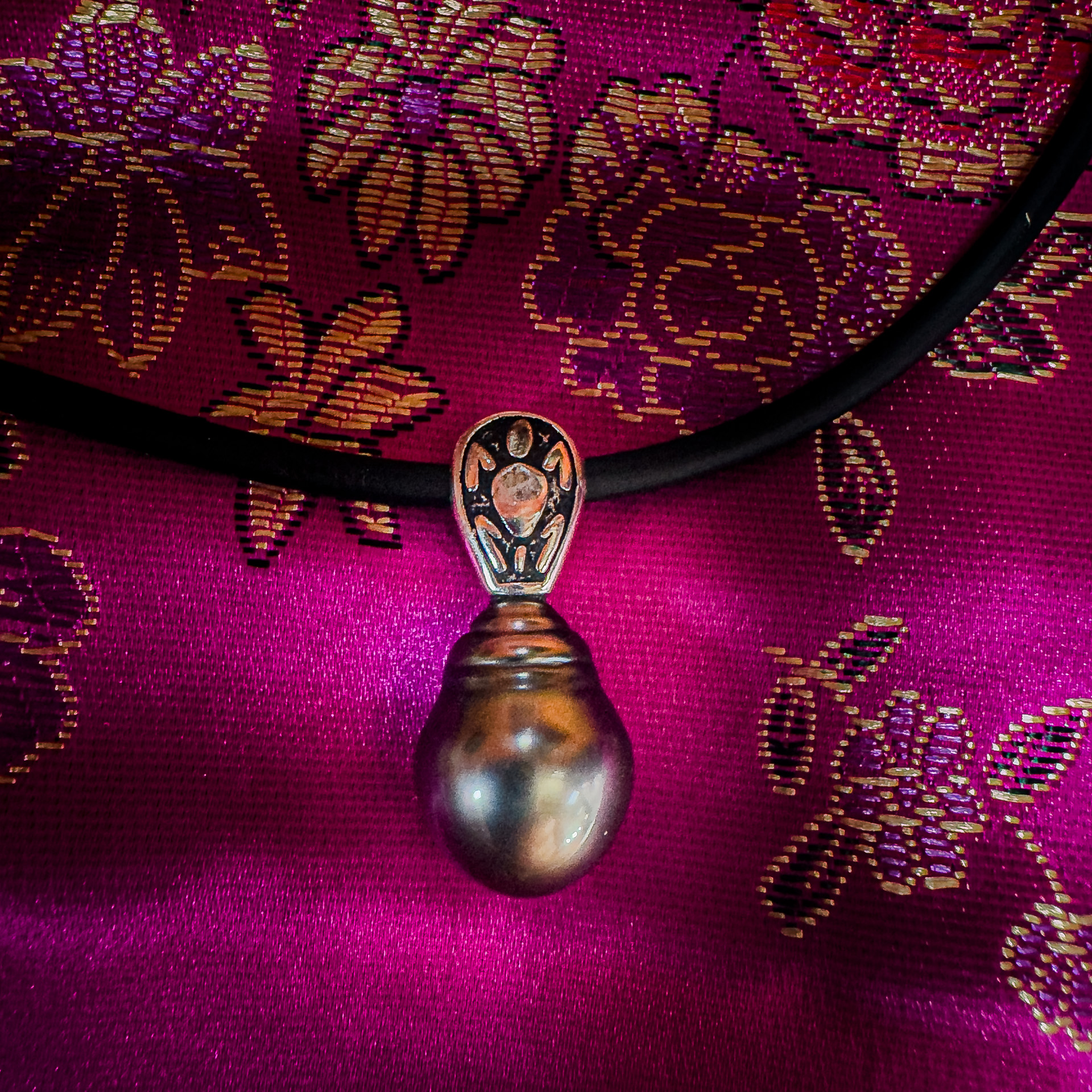 A pendant with a dark pearl and ornate metal design on a black cord, set against vibrant pink fabric with gold floral patterns.