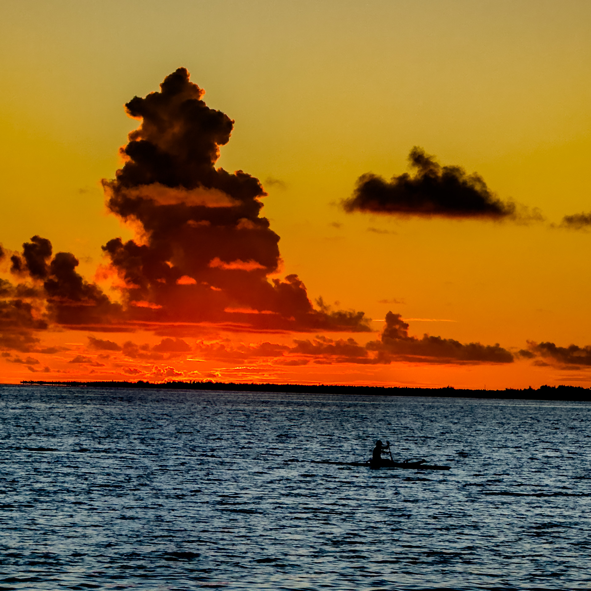 A person kayaking on a calm ocean at sunset, with vibrant orange and yellow hues in the sky and dramatic clouds on the horizon.