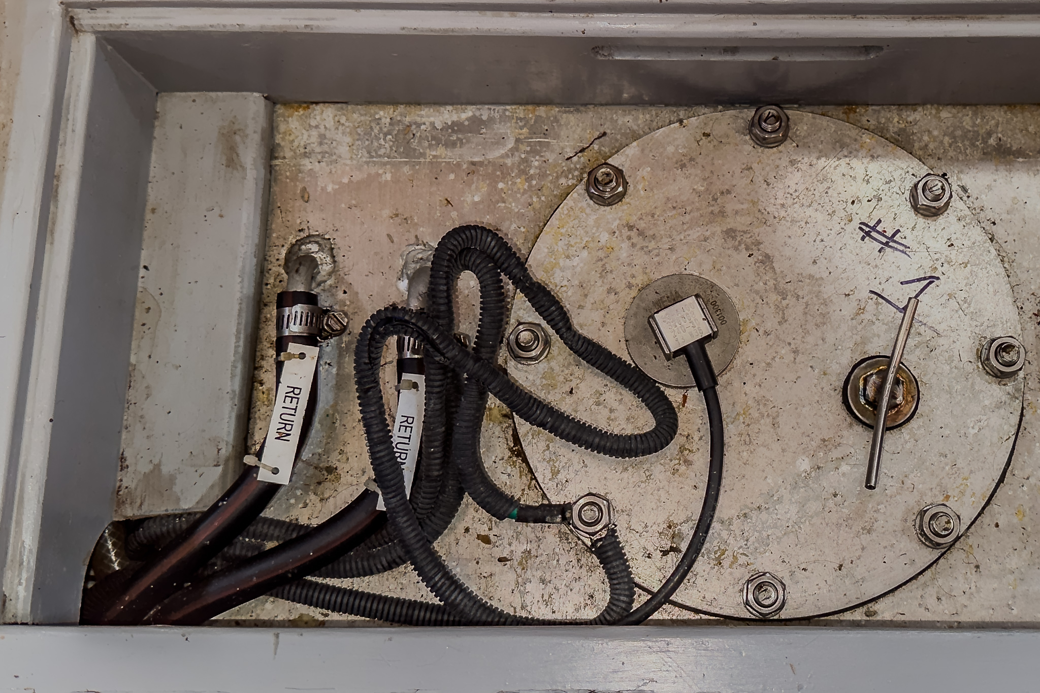 SV Wanderlust's stainless-steel center fuel tank with a round, bolted inspection plate, labeled cables, and a small gauge on a weathered surface.