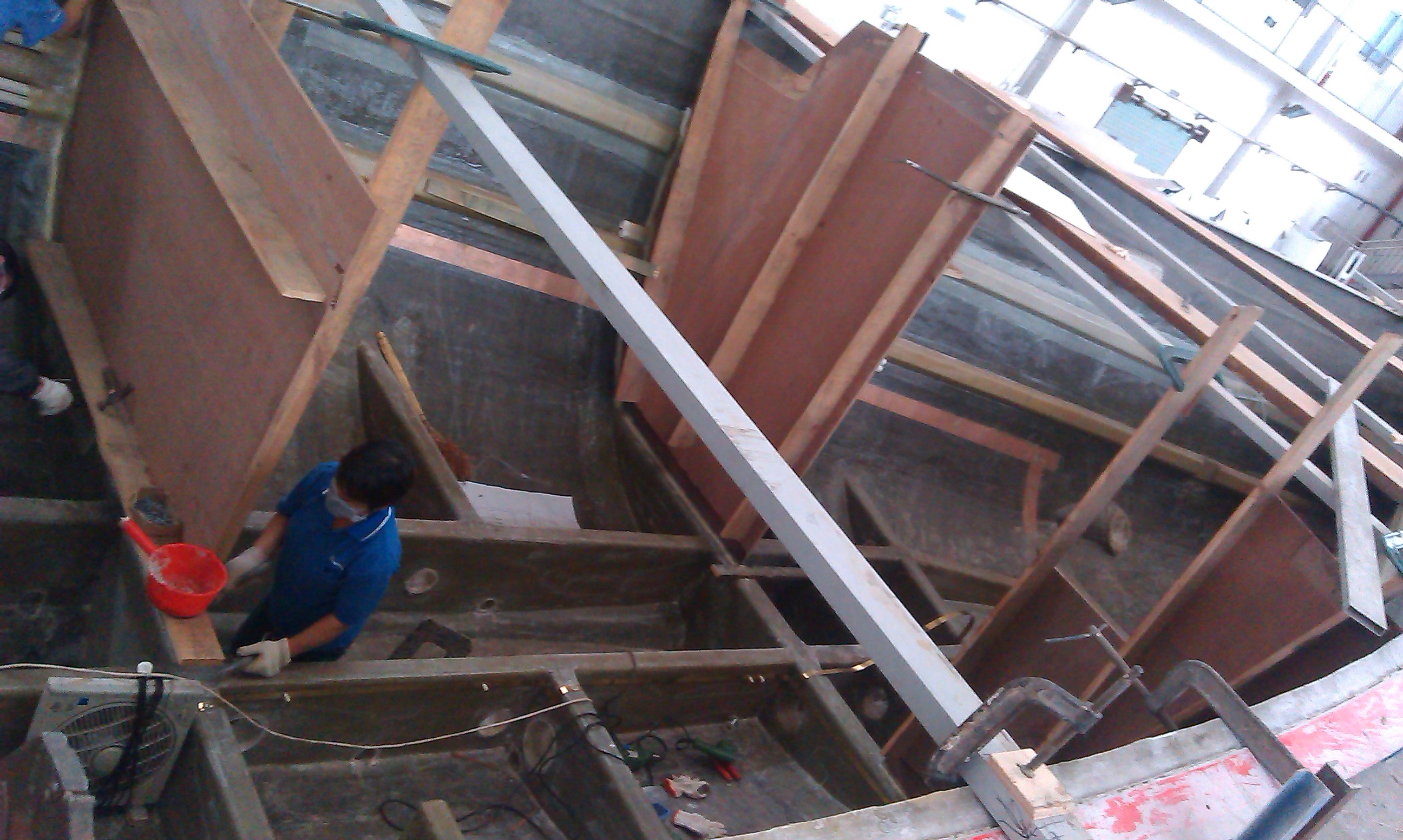 Person working on a large sailboat hull under construction, with wooden frames and metal supports inside a workshop.