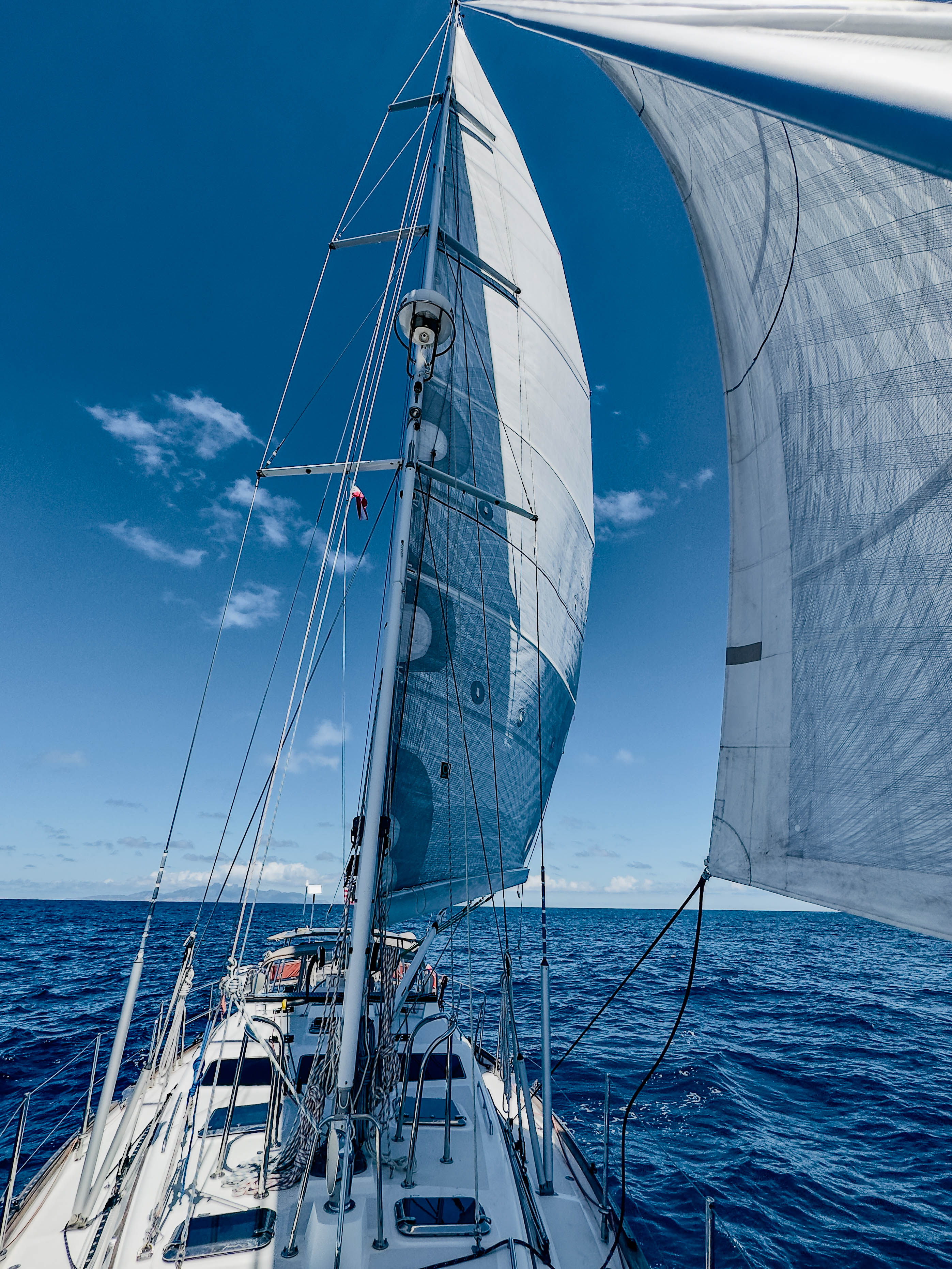 View from a Wanderlust's foredeck aft to tall mast and billowing white sails over deep blue ocean under a clear sky.