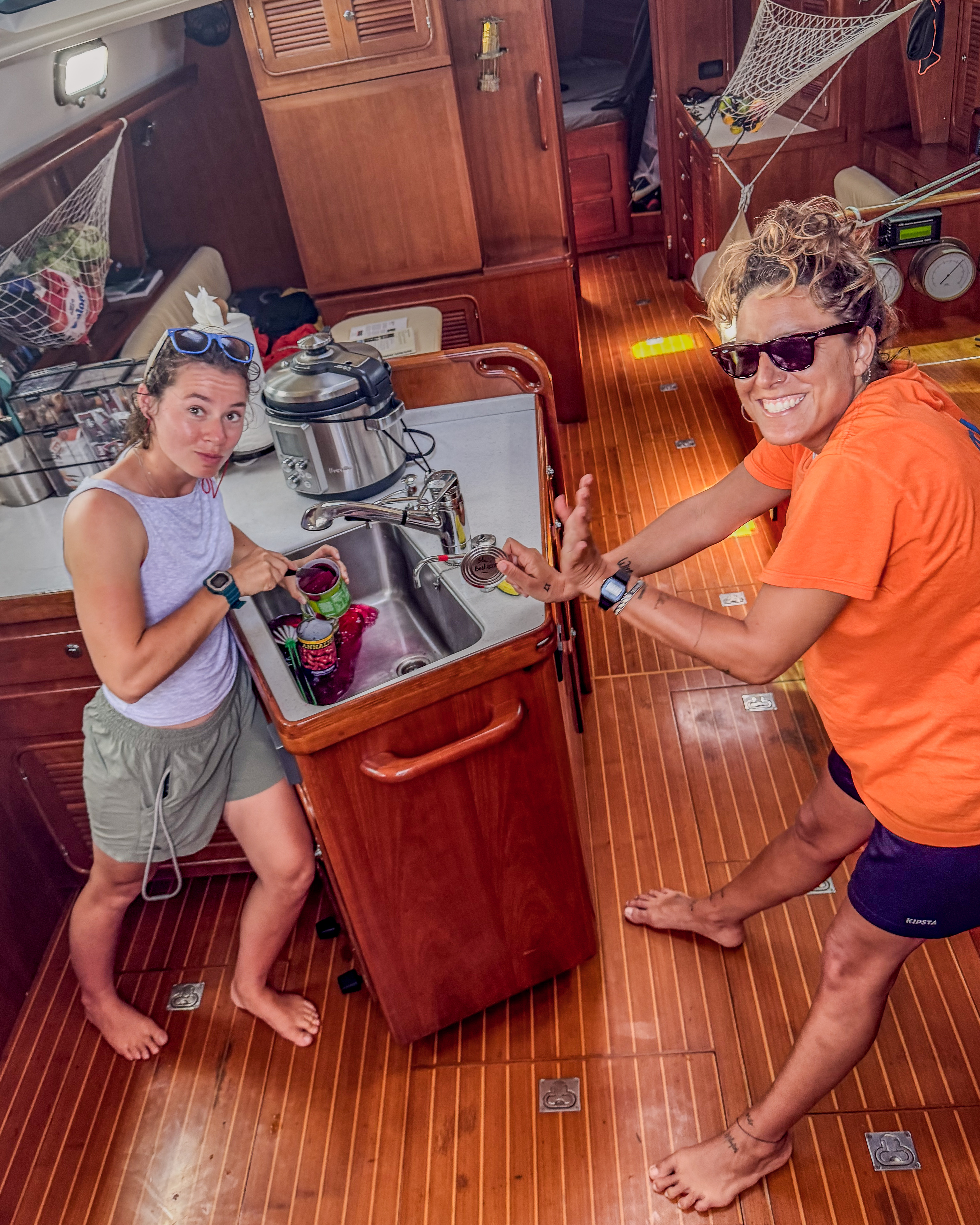 Two people smiling in a boat galley, preparing food at the sink. The wooden floor and cabinets create a warm ambiance.