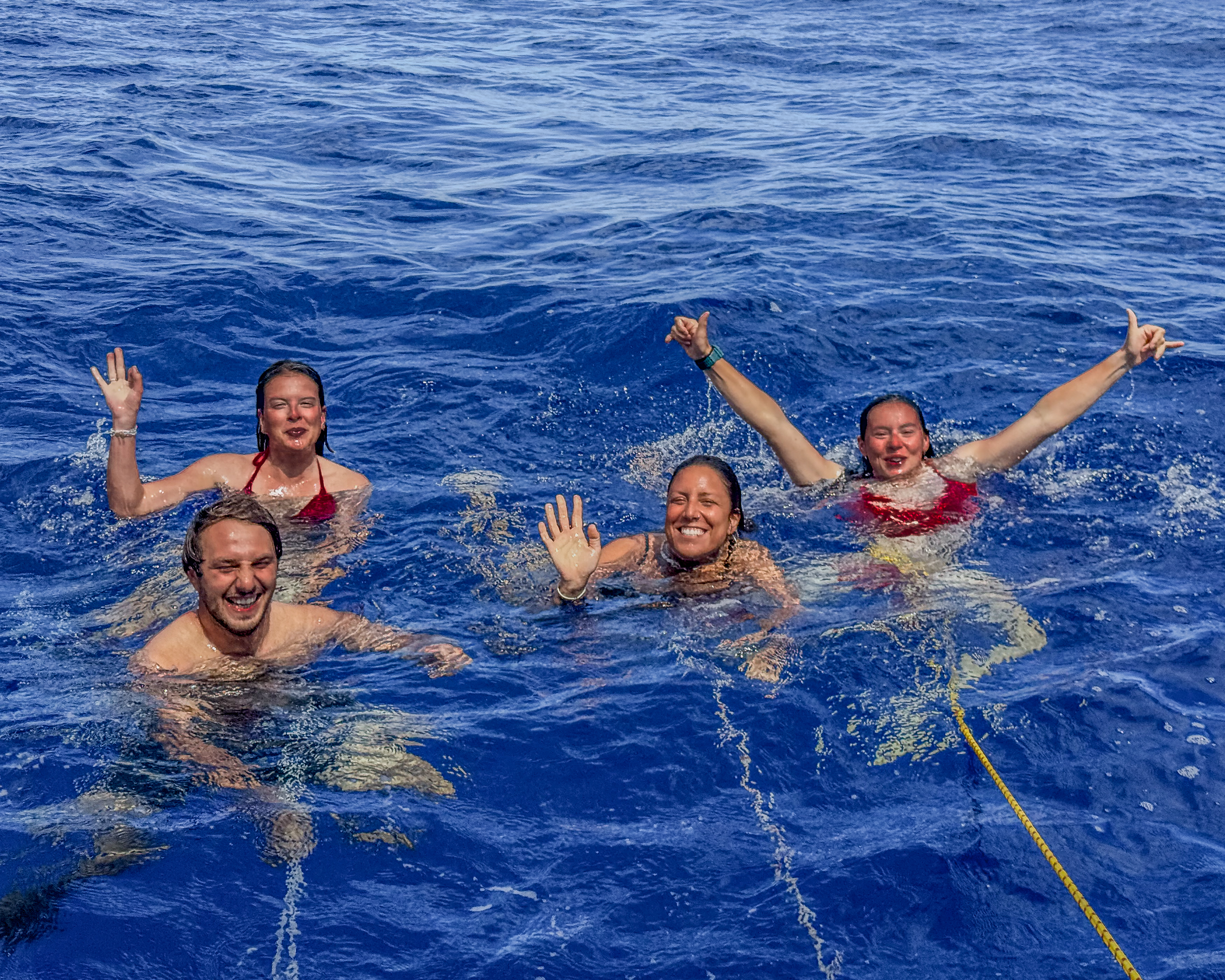 Four people smiling and waving while swimming in deep blue ocean water, holding onto a rope.