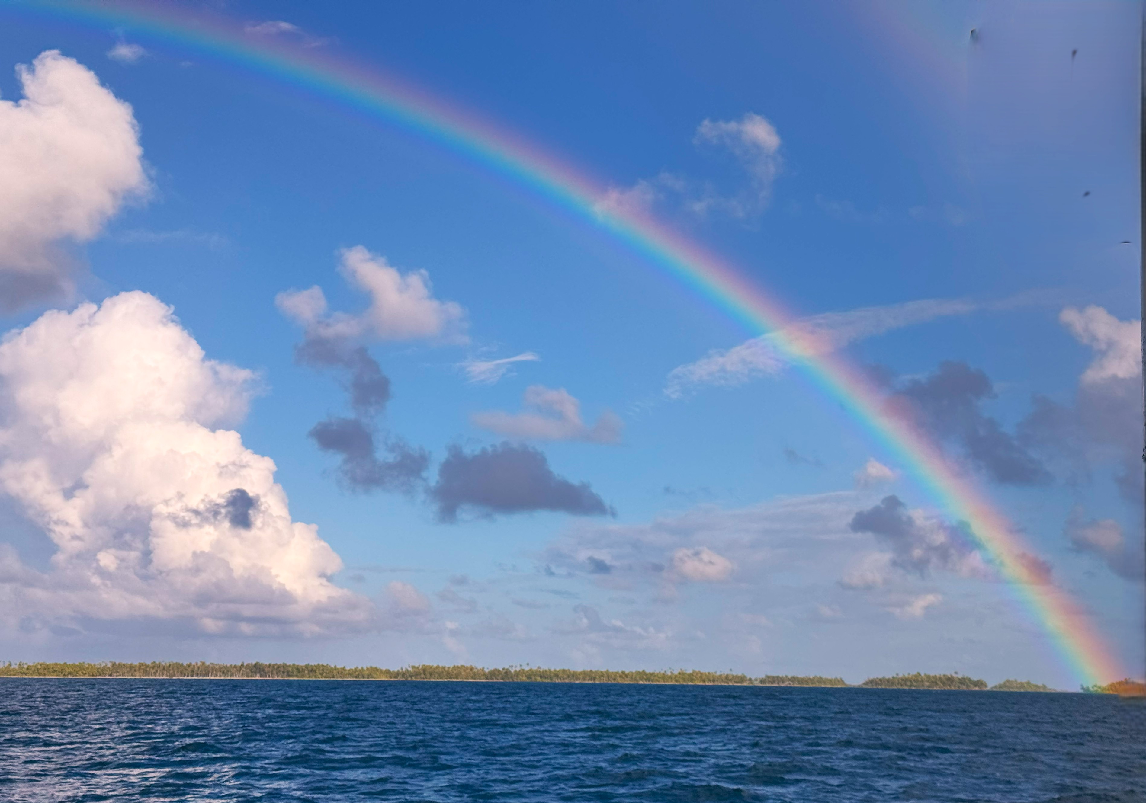 A vibrant rainbow arcs across a blue sky with fluffy clouds, above a calm ocean and distant tree-lined horizon, as seen from sailing vessel Wanderlust.