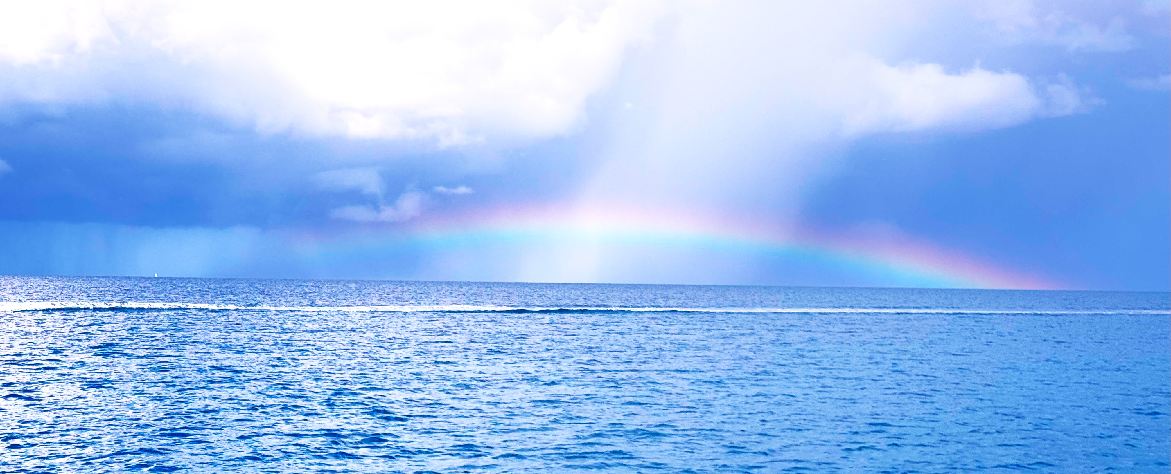 A vibrant rainbow arcs over the Rangiroa lagoon under a cloudy sky, with sunlight illuminating the water's surface.