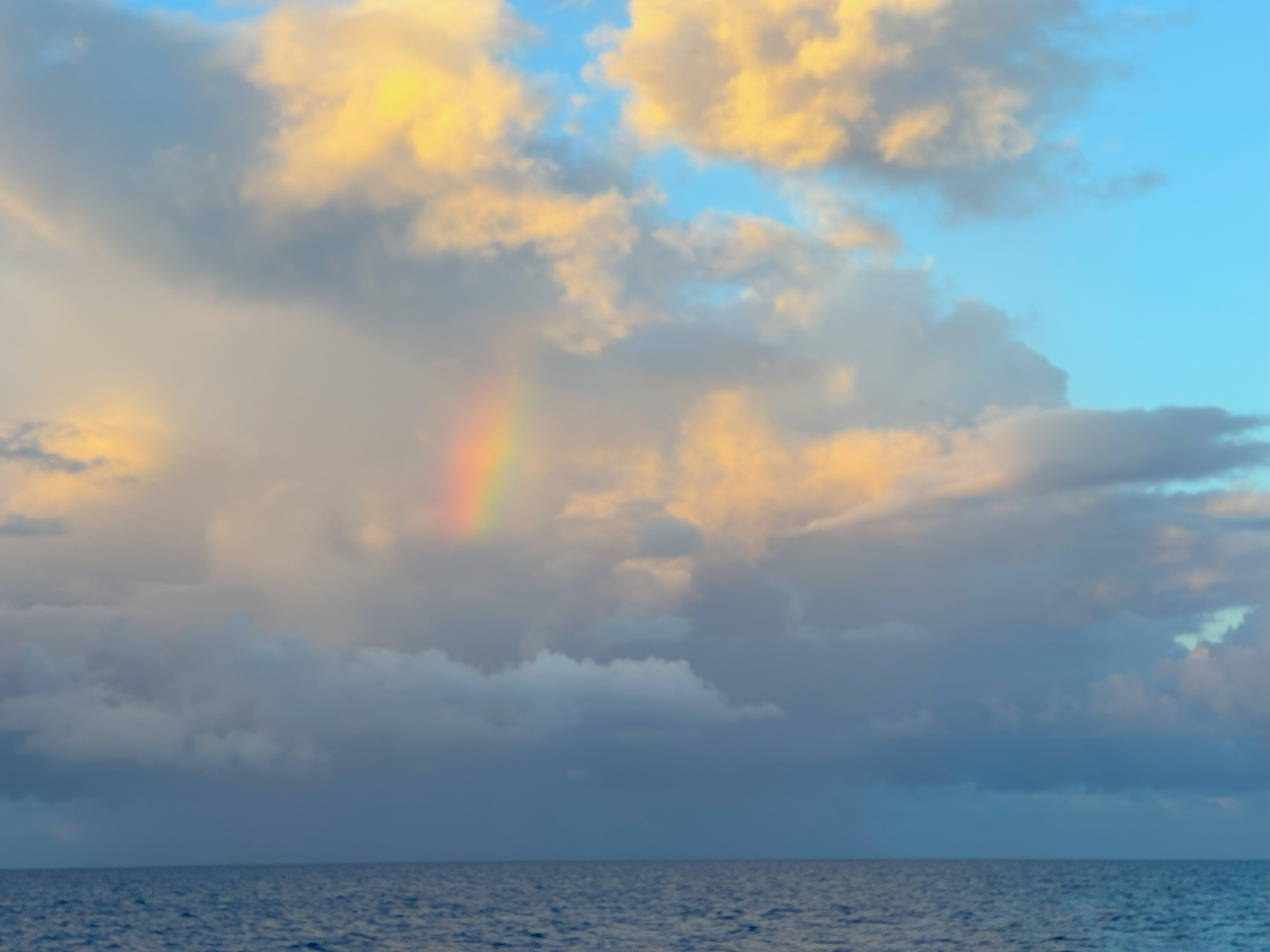 A faint rainbow appears among fluffy clouds over a calm Rangiroa lagoon, with a blue sky and golden sunlight illuminating the scene.