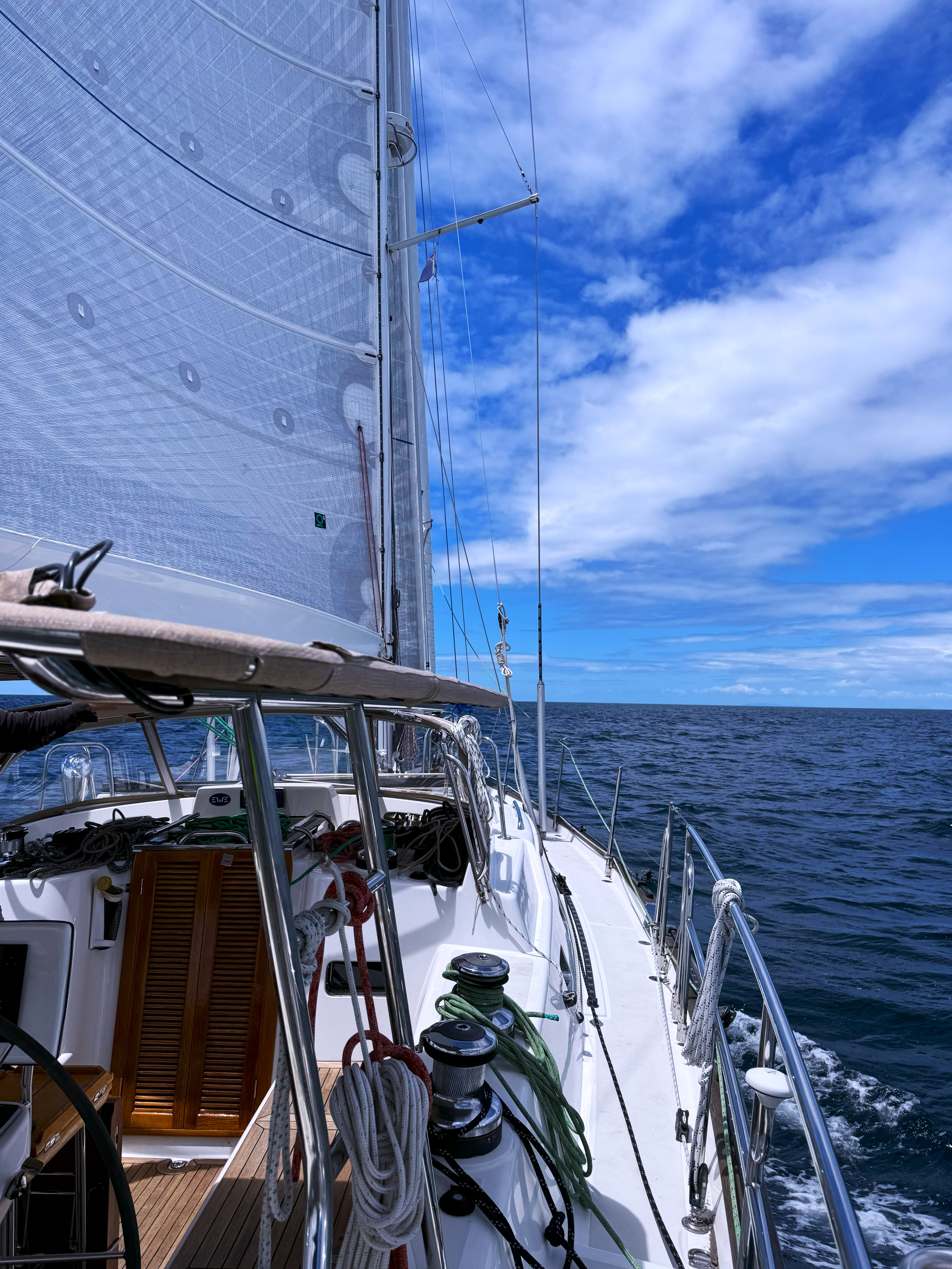 Wanderlust, a sailboat on open sea with white sails against a partly cloudy blue sky, view from the deck showing ropes and equipment.