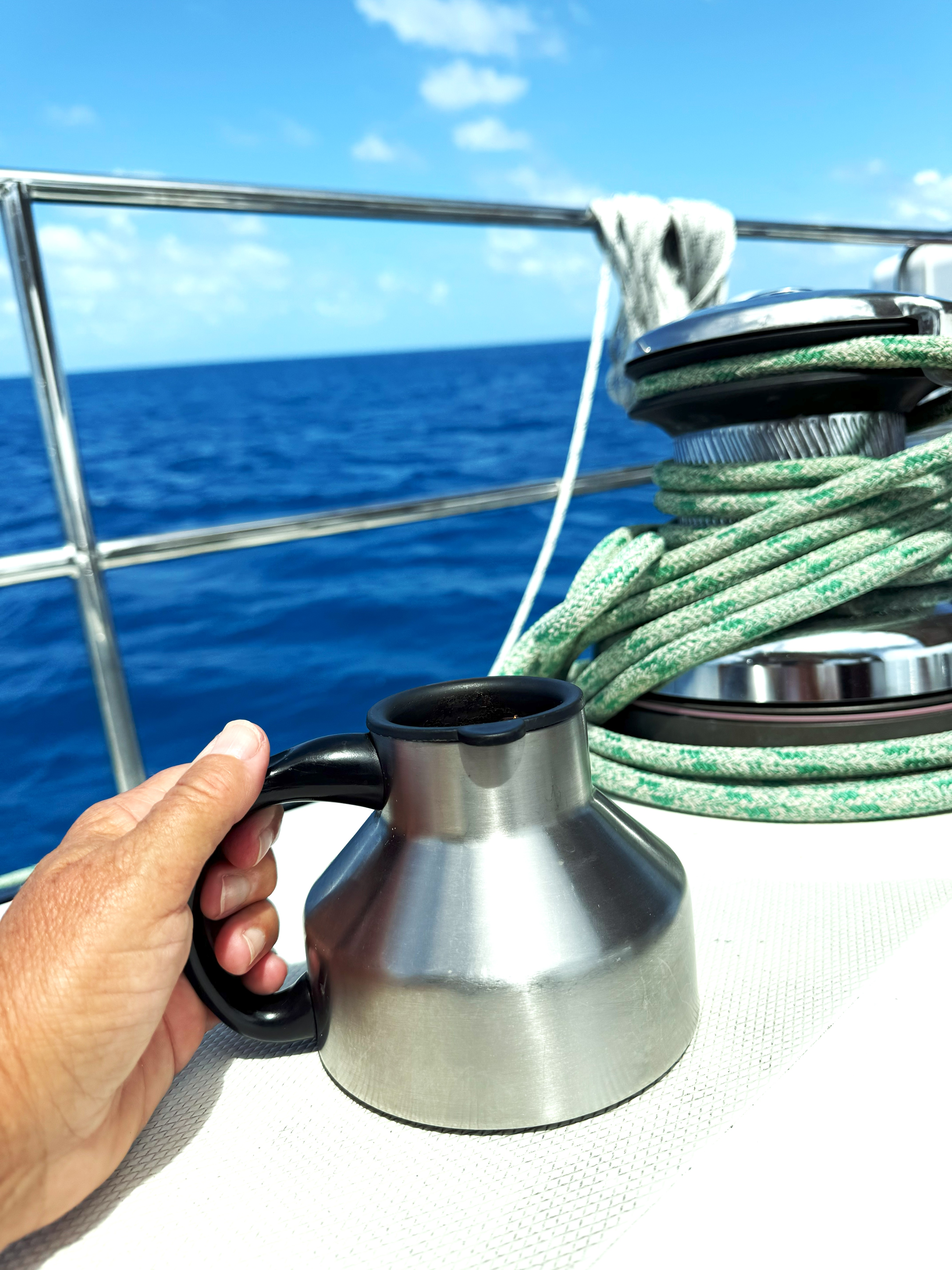 Hand holding a metal coffee mug on a sailboat deck with coiled green line, against a backdrop of blue ocean and sky.