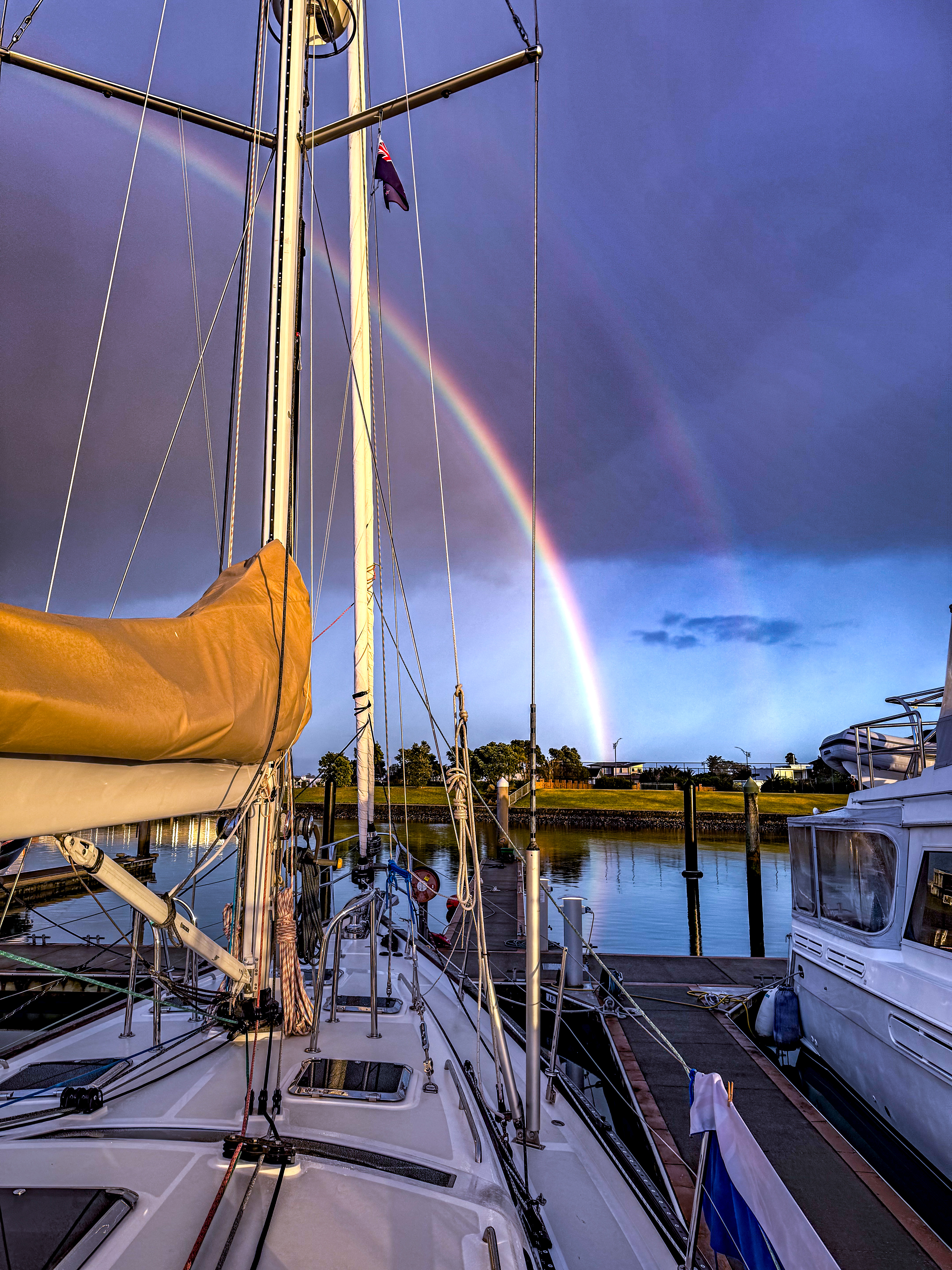 Sailboat Wanderlust docked in Marsden Cove Marina with a vivid double rainbow in the cloudy sky, reflecting on the calm morning water.