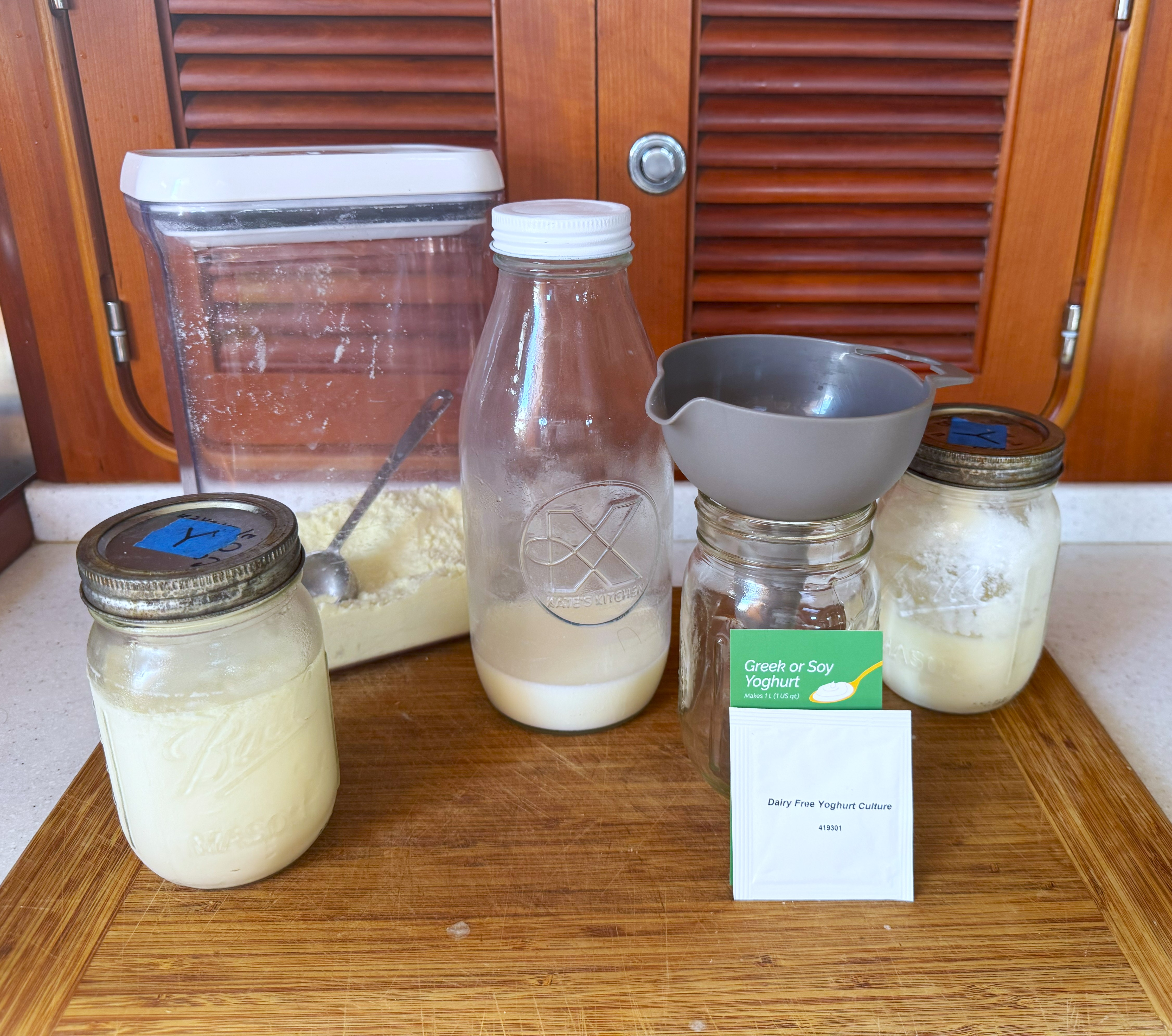 Ingredients and tools for making yogurt on a wooden board, including jars, a bottle, a container of powder, and a sachet labeled "Greek or Soy Yogurt."