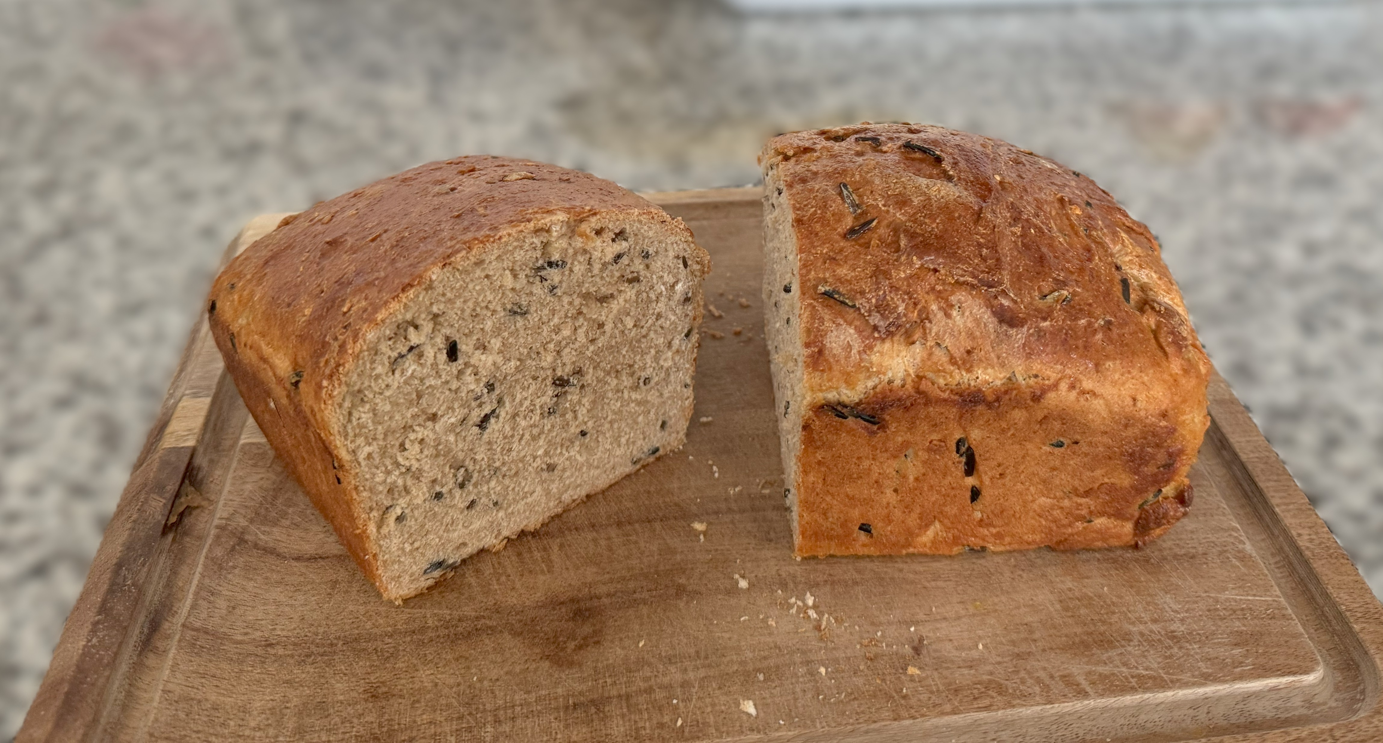 Sliced loaf of homemade bread with visible seeds on a wooden cutting board, set on a speckled countertop.