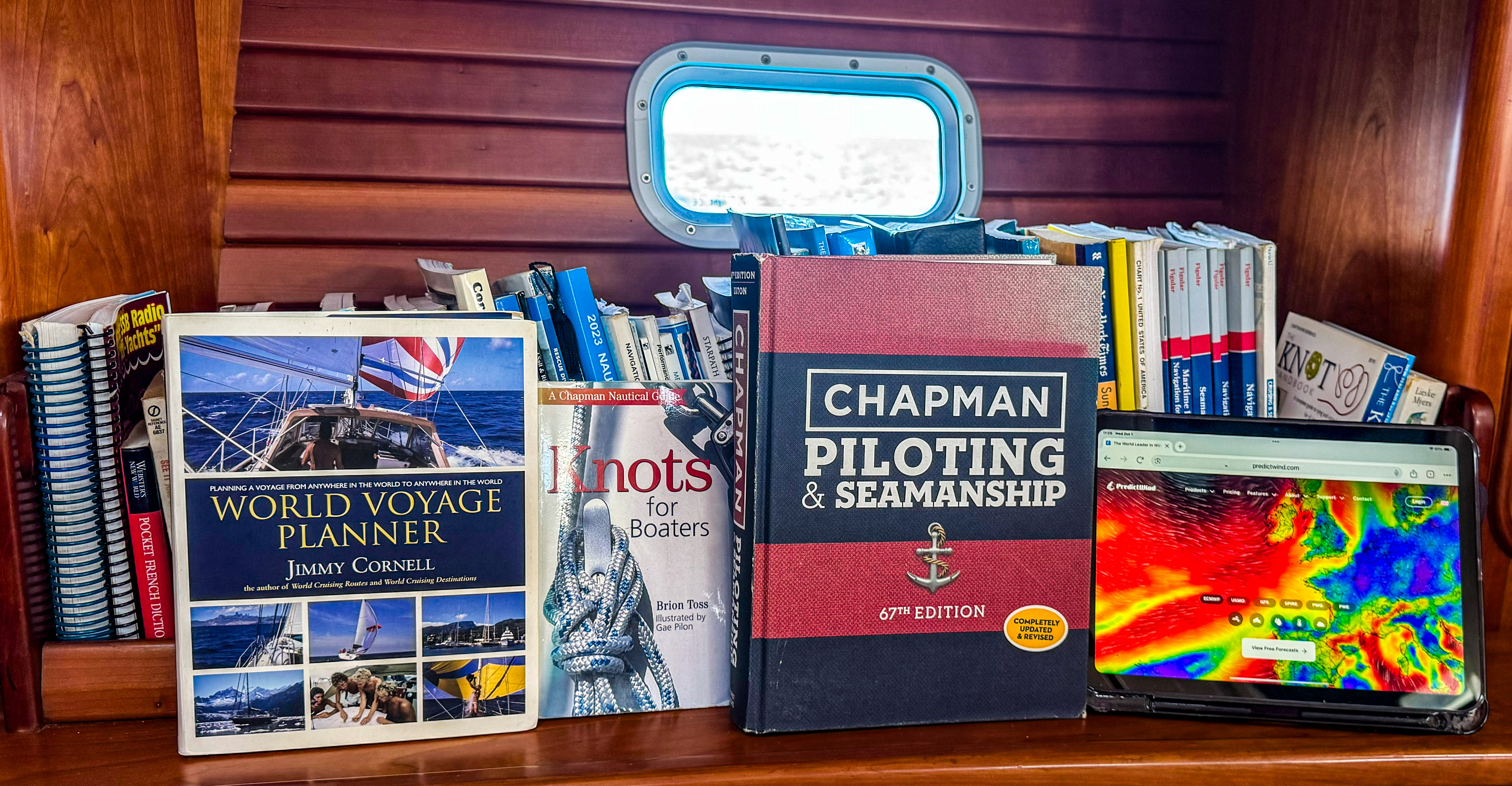 Books on sailing, including "World Voyage Planner" and "Chapman Piloting & Seamanship," are displayed on a wooden shelf with a tablet.