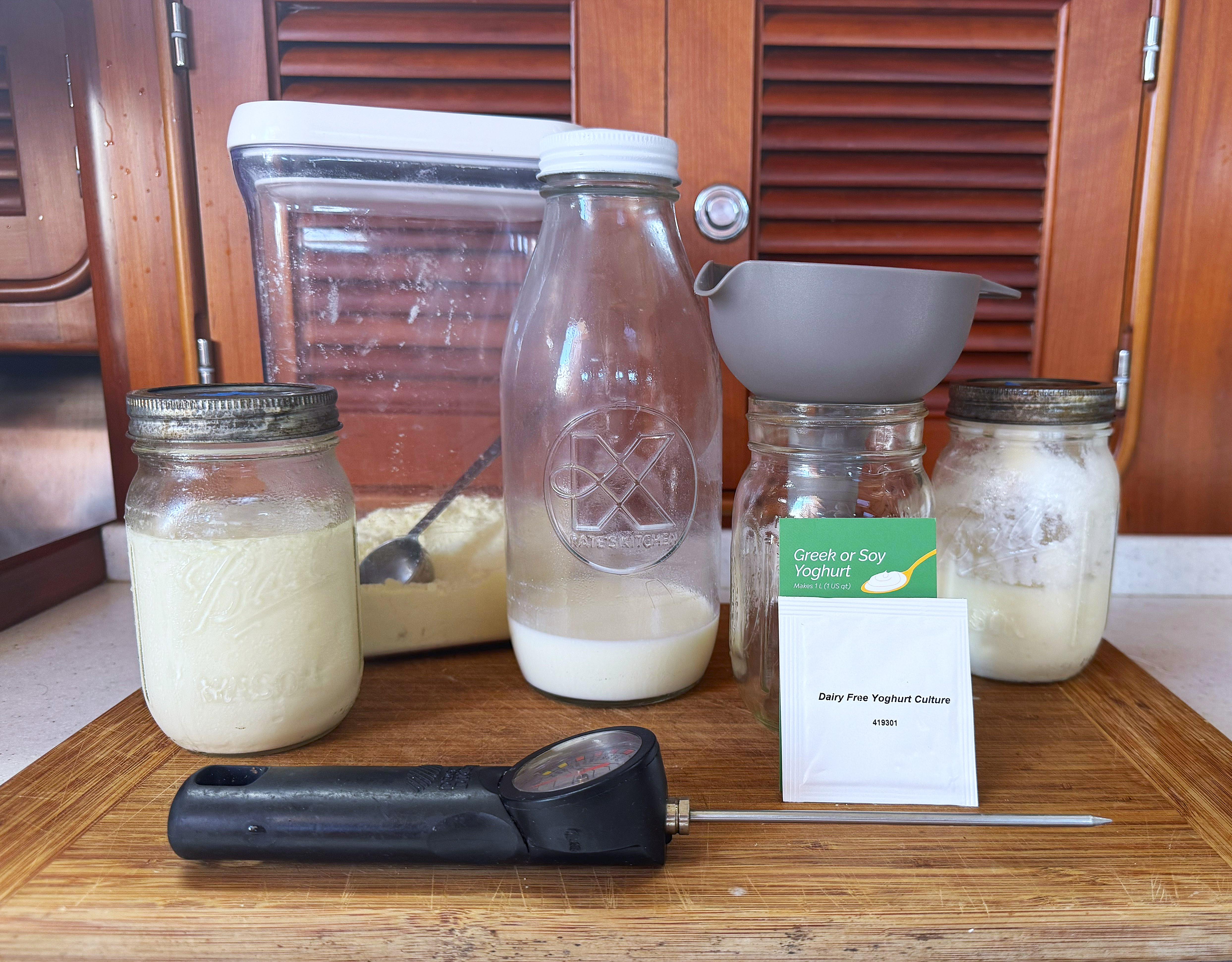 Homemade yogurt setup with jars, a glass bottle, a thermometer, a strainer, and a packet labeled "Dairy Free Yogurt Culture" on a wooden board.