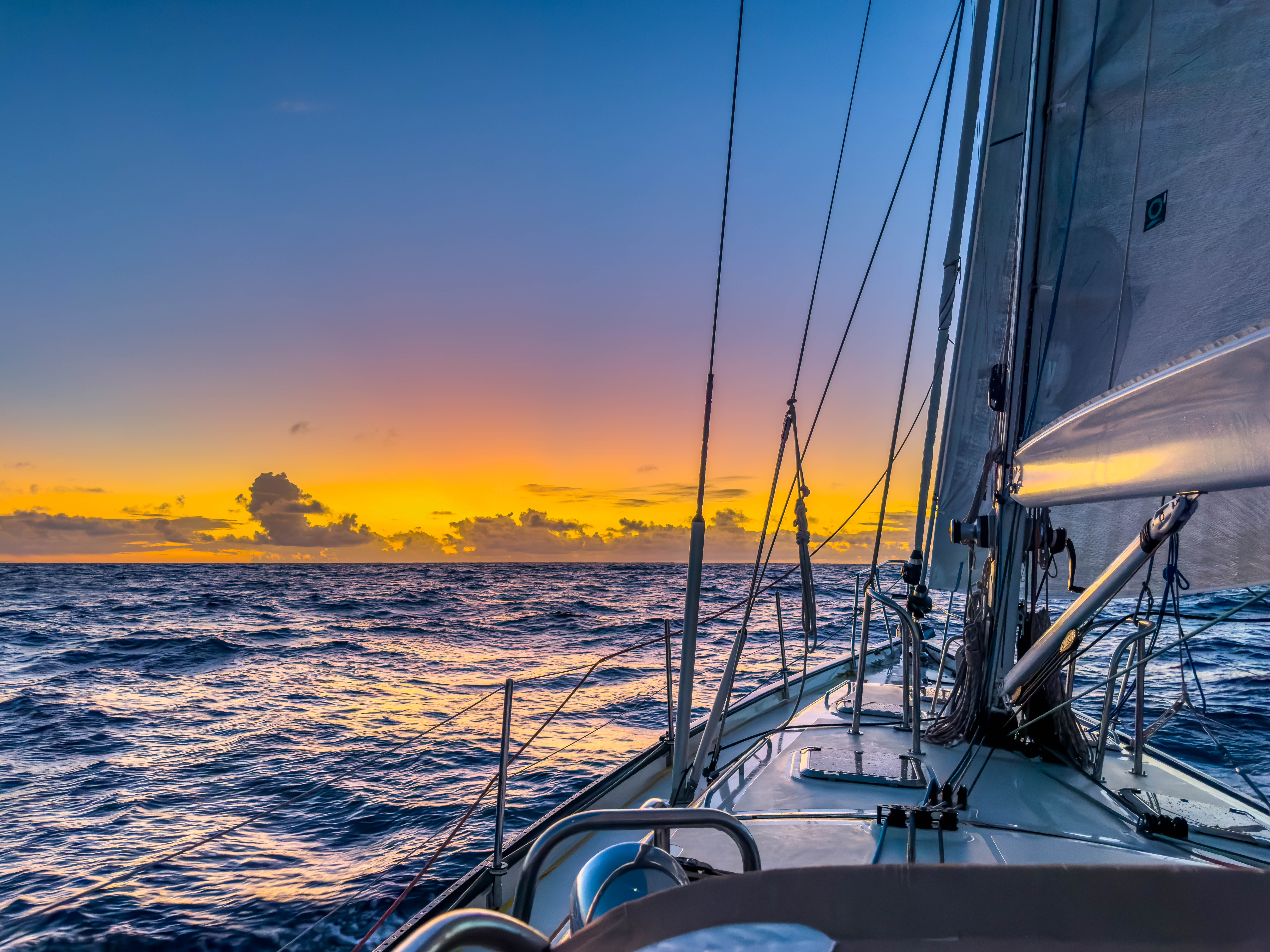 Sailing vessel Wanderlust glides on calm ocean waters at sunset, with vibrant orange and blue skies reflected on the sea.
