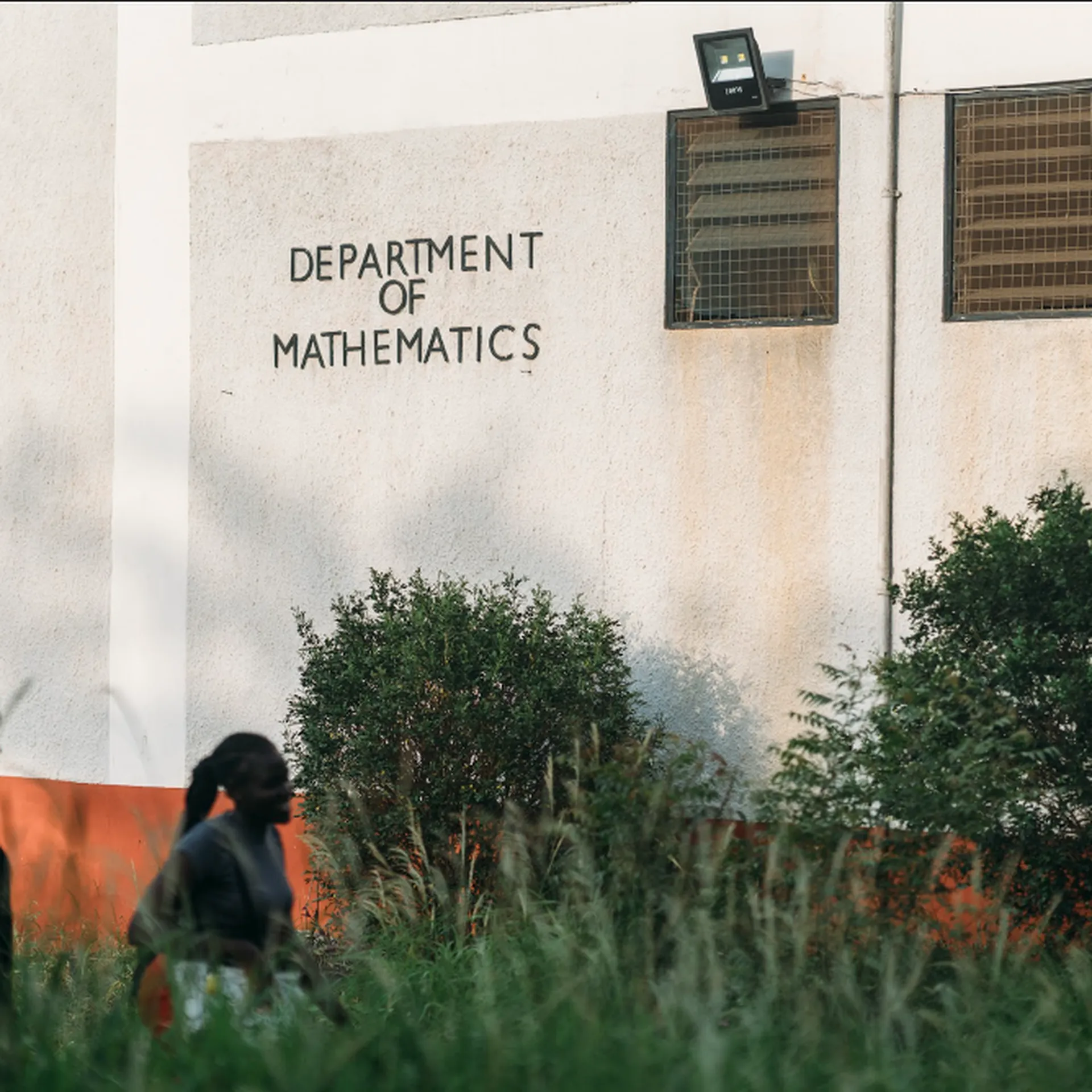 Two people walk past a building with "Department of Mathematics" on the wall surrounded by greenery.