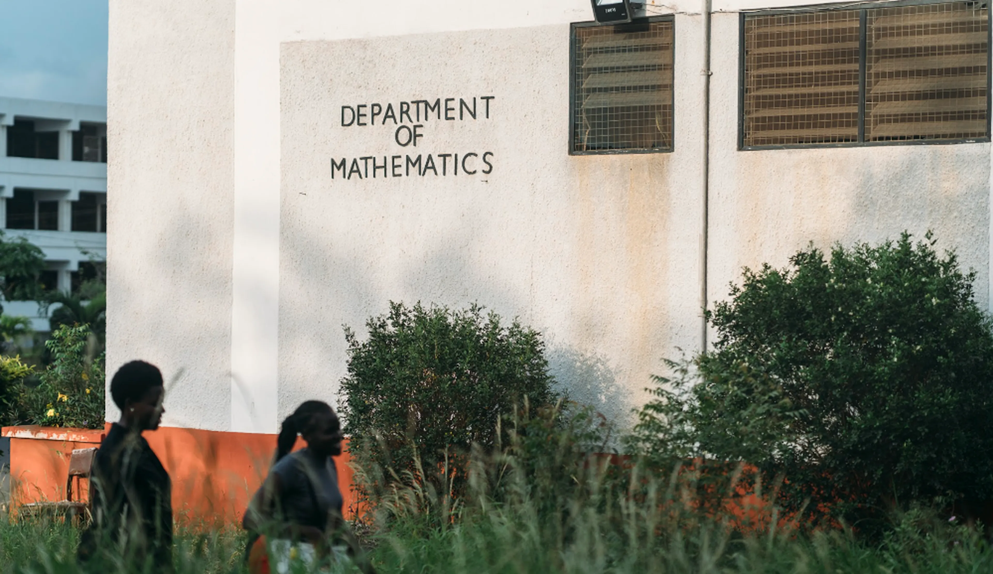 Two people walk past a building with "Department of Mathematics" on the wall surrounded by greenery.