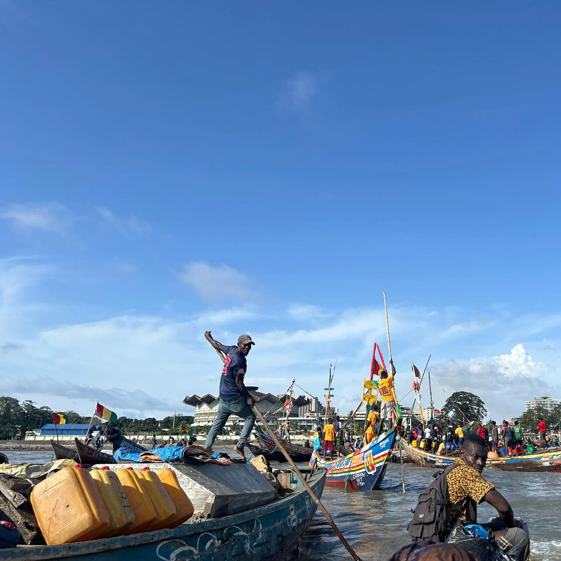 Fishermen on colorful boats at a busy shoreline, with clear blue skies and distant trees in the background.