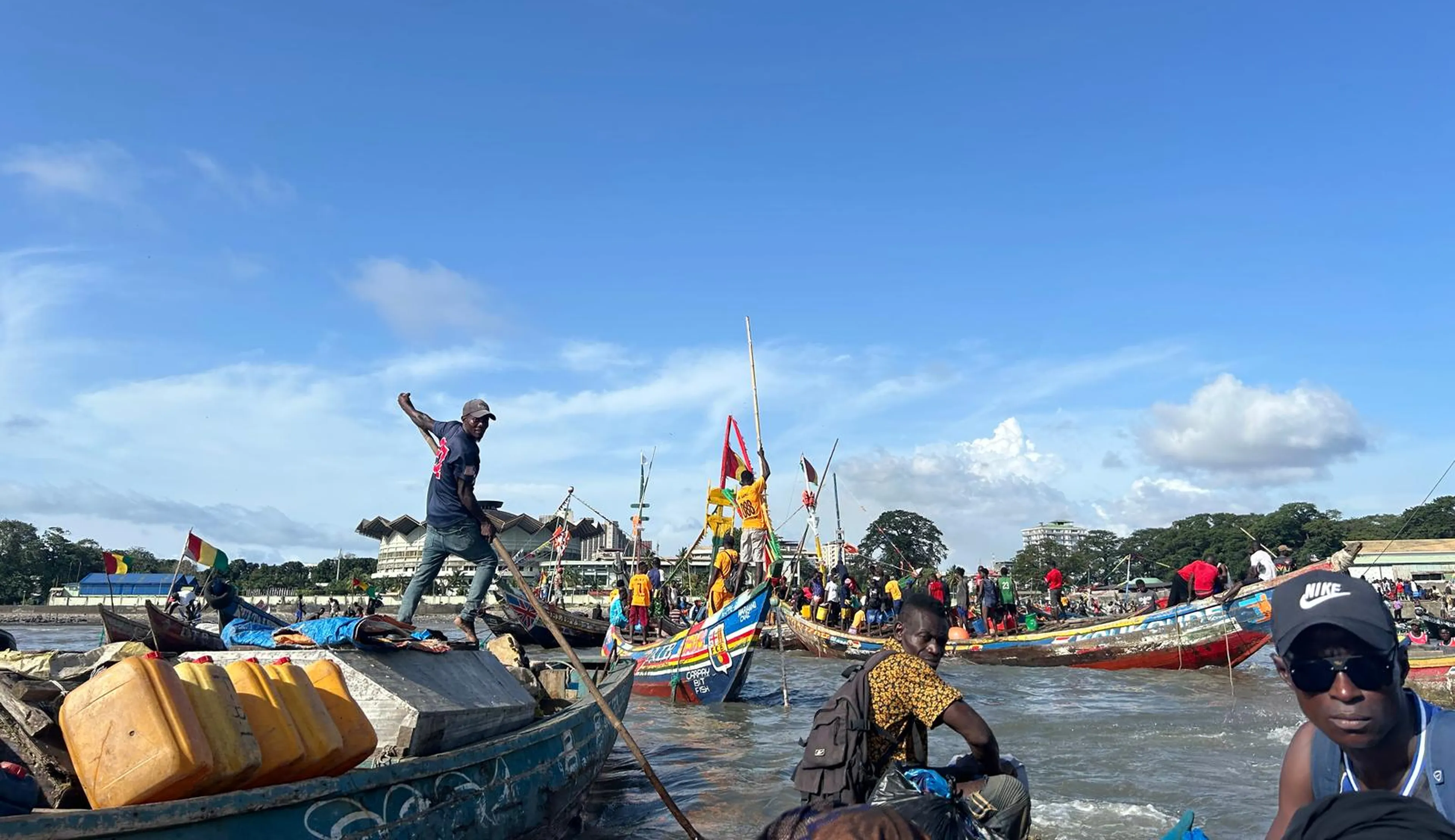 Fishermen on colorful boats at a busy shoreline, with clear blue skies and distant trees in the background.
