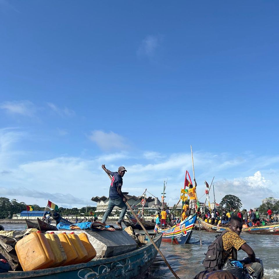 Fishermen on colorful boats at a busy shoreline, with clear blue skies and distant trees in the background.