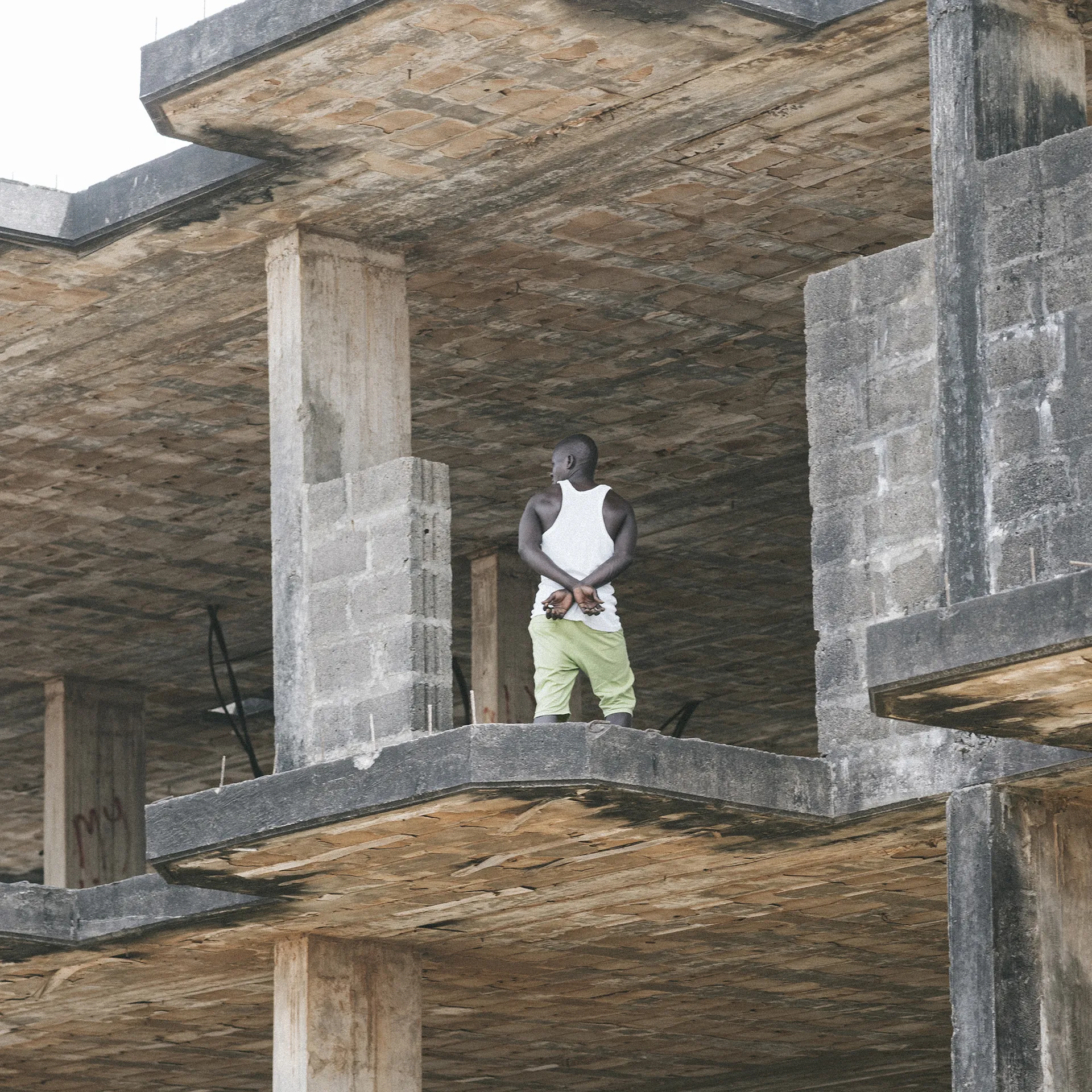 Man in white tank top and green pants stands on an unfinished concrete building floor, gazing outward.