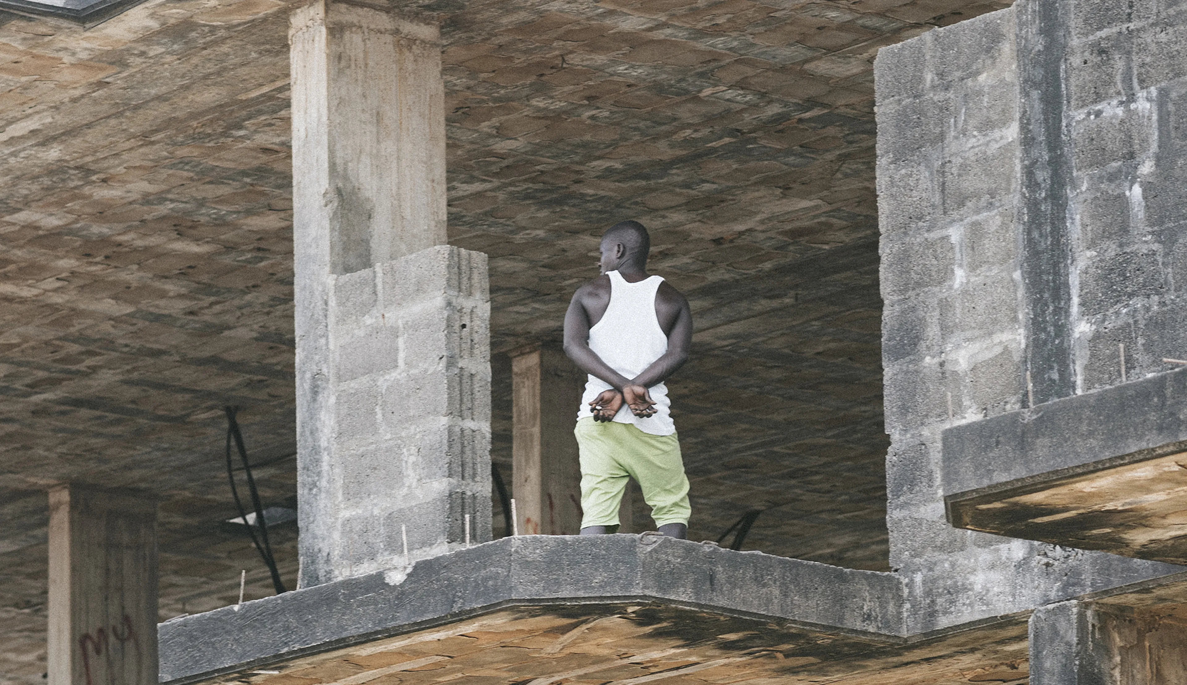 Man in white tank top and green pants stands on an unfinished concrete building floor, gazing outward.