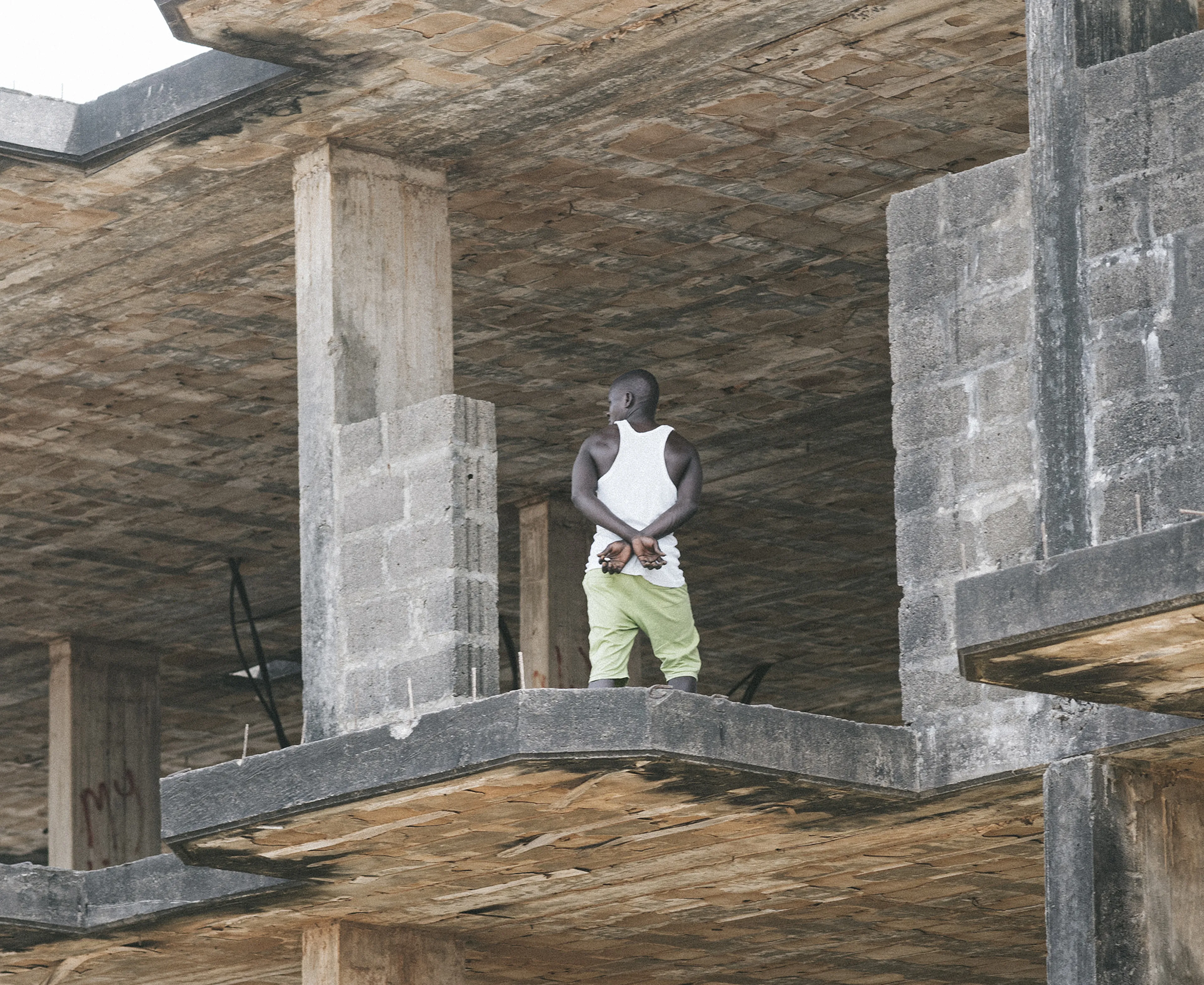 Man in white tank top and green pants stands on an unfinished concrete building floor, gazing outward.