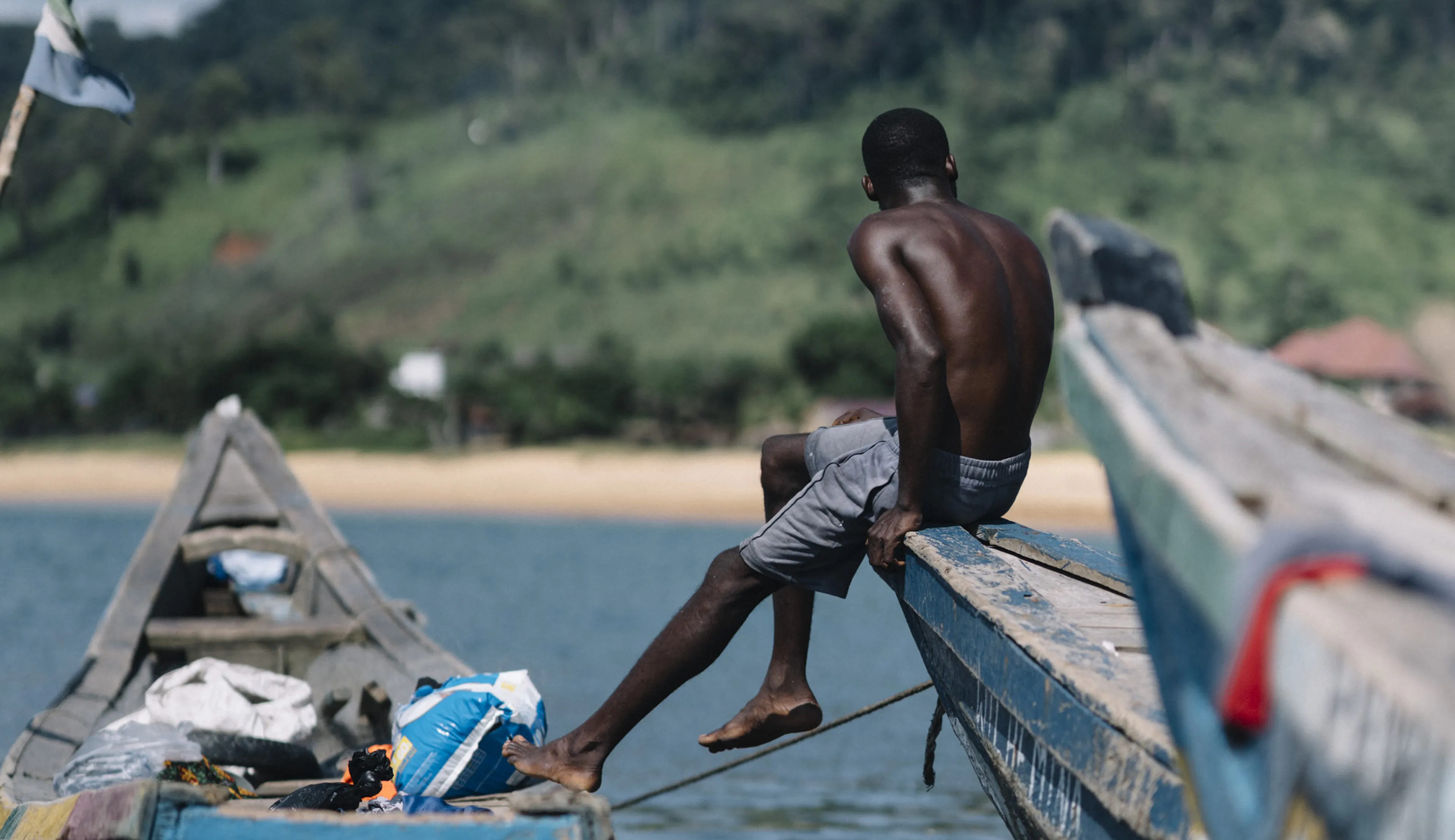 A man sits on the edge of a colorful wooden boat on a calm lake, with a lush green hill in the background under a clear blue sky.