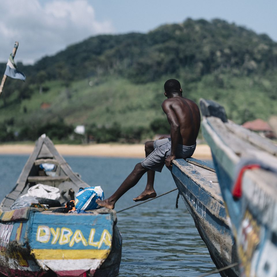 A man sits on the edge of a colorful wooden boat on a calm lake, with a lush green hill in the background under a clear blue sky.