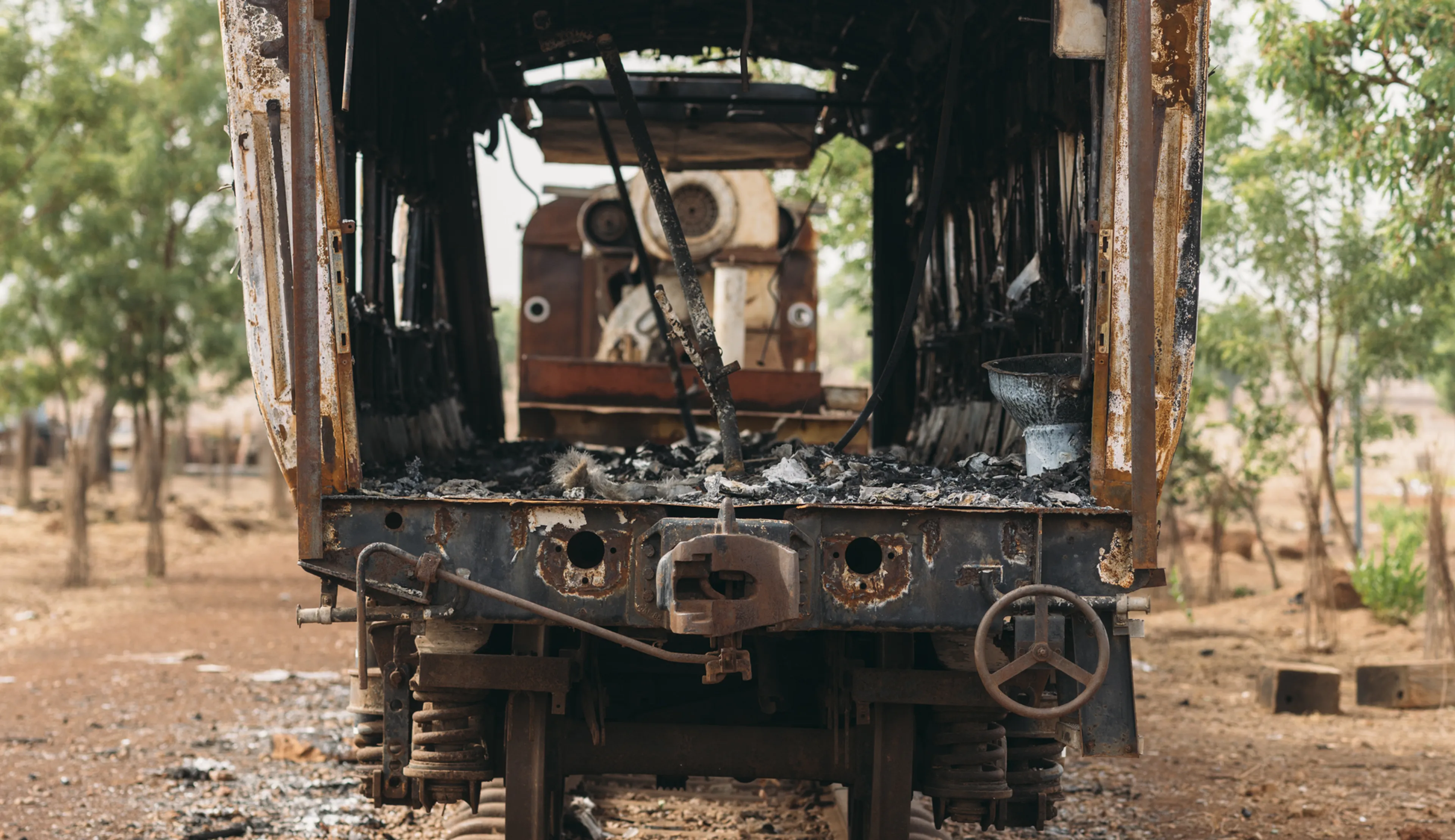 Front view of an abandoned, rusted train carriage on old tracks in a barren landscape with scattered trees in the background.
