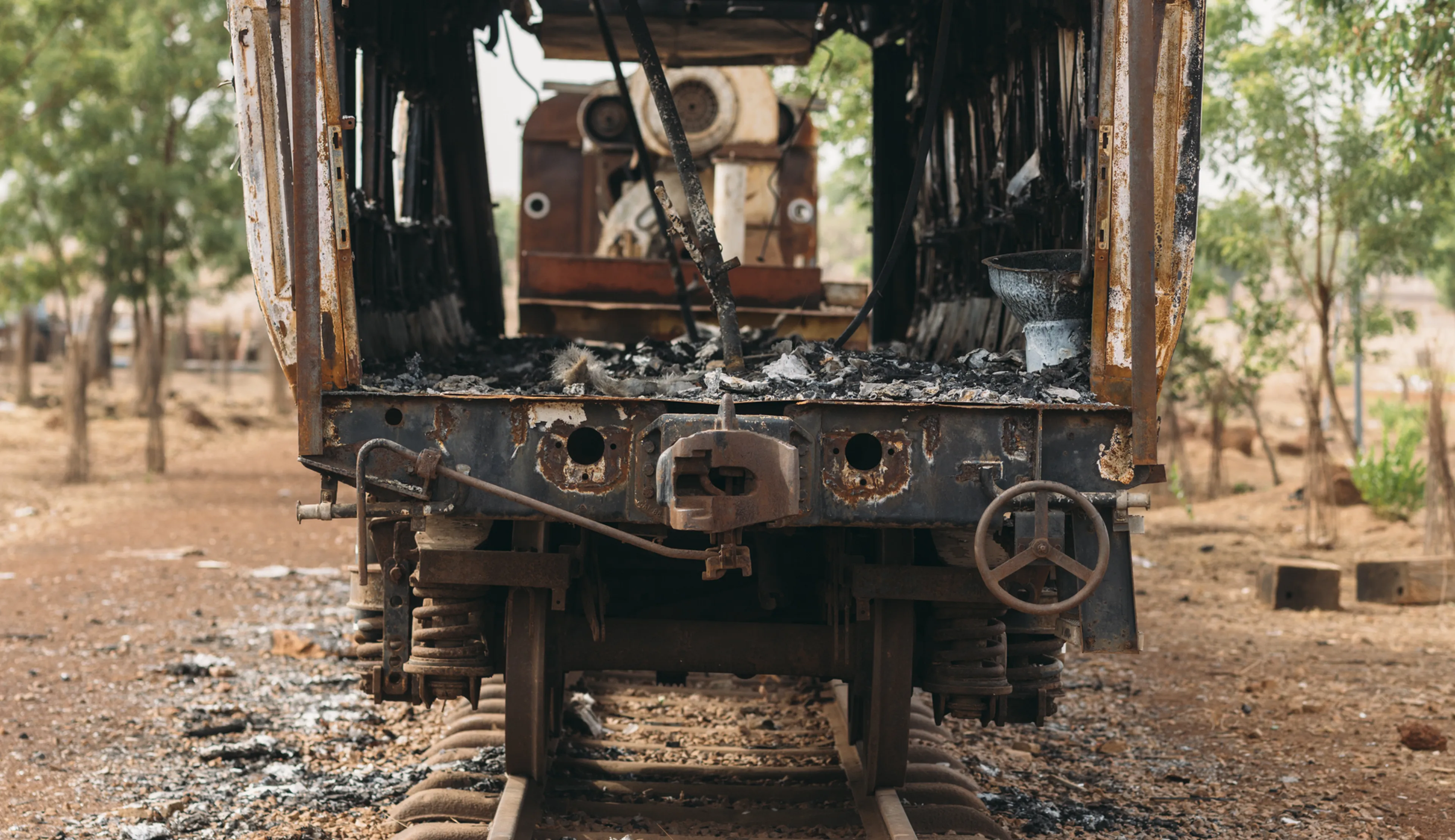 Front view of an abandoned, rusted train carriage on old tracks in a barren landscape with scattered trees in the background.