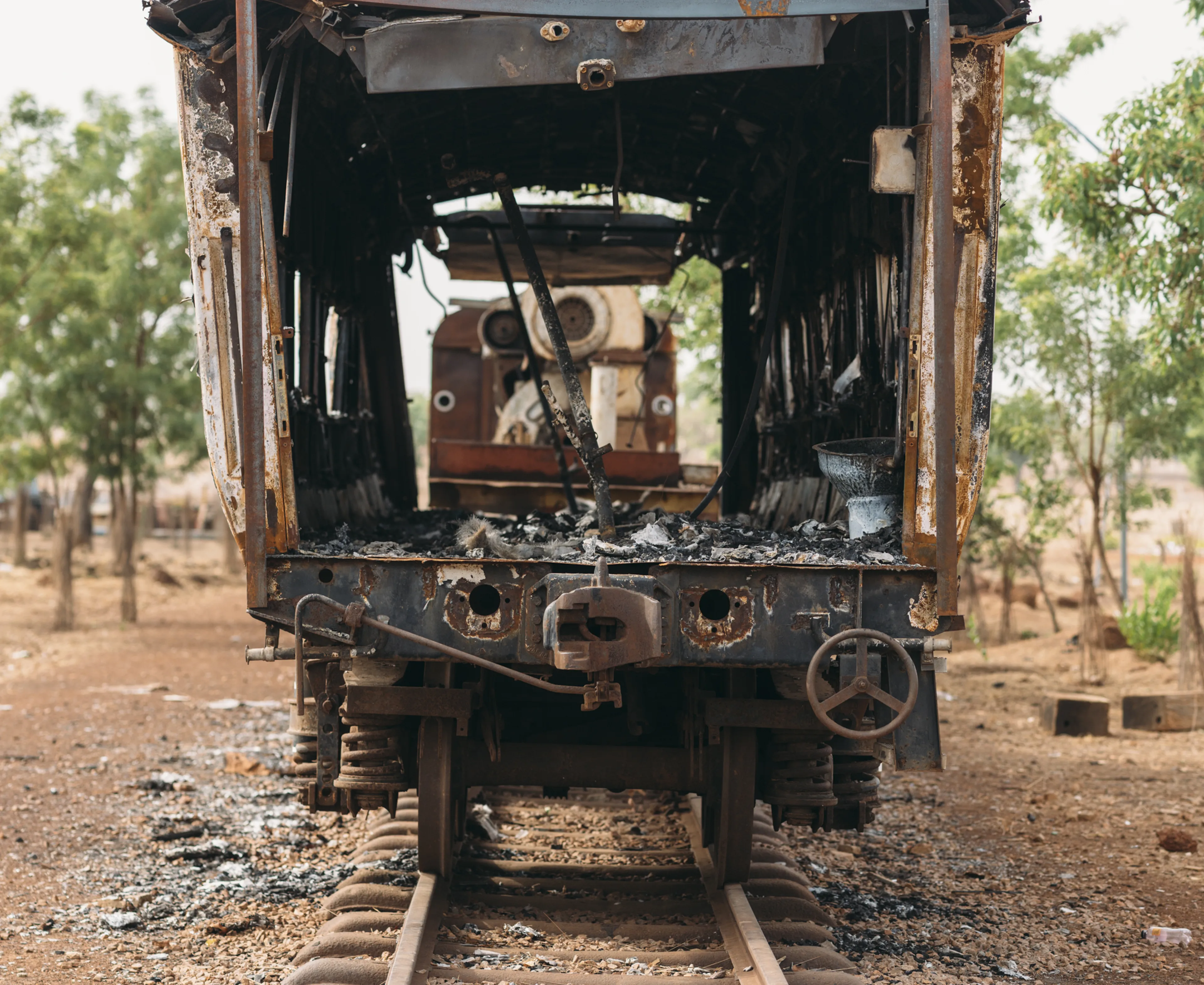 Front view of an abandoned, rusted train carriage on old tracks in a barren landscape with scattered trees in the background.