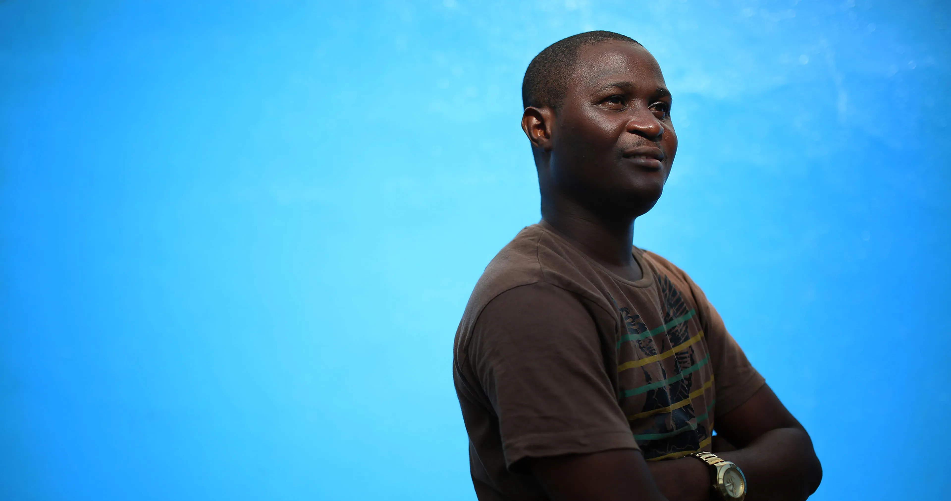 Person in a brown shirt with crossed arms, standing against a bright blue background, looking confidently to the side.