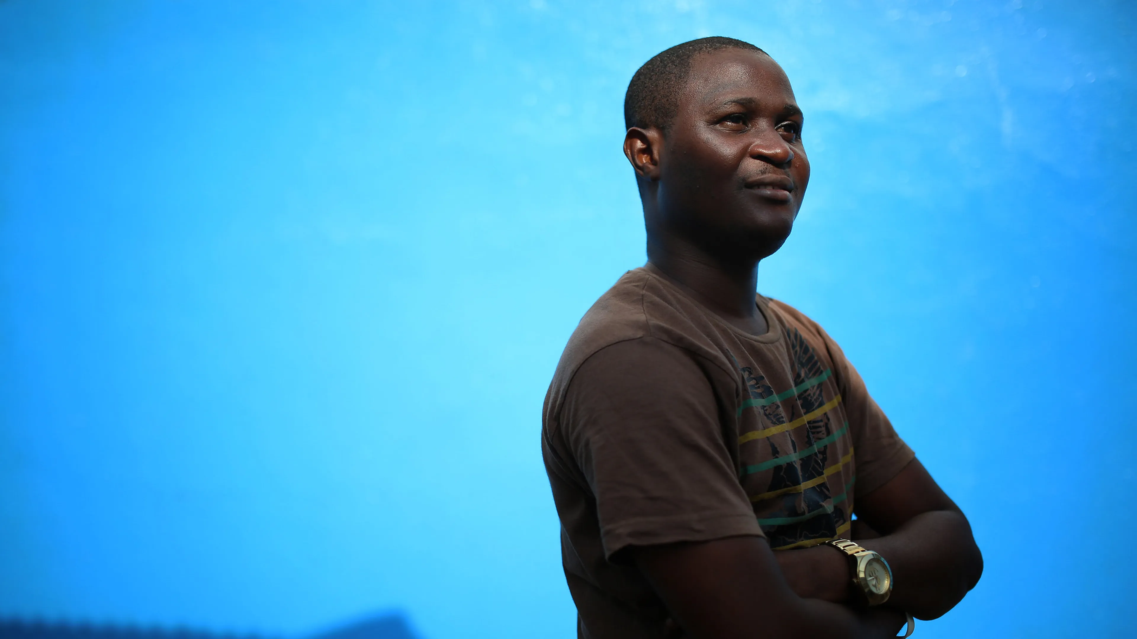 Person in a brown shirt with crossed arms, standing against a bright blue background, looking confidently to the side.
